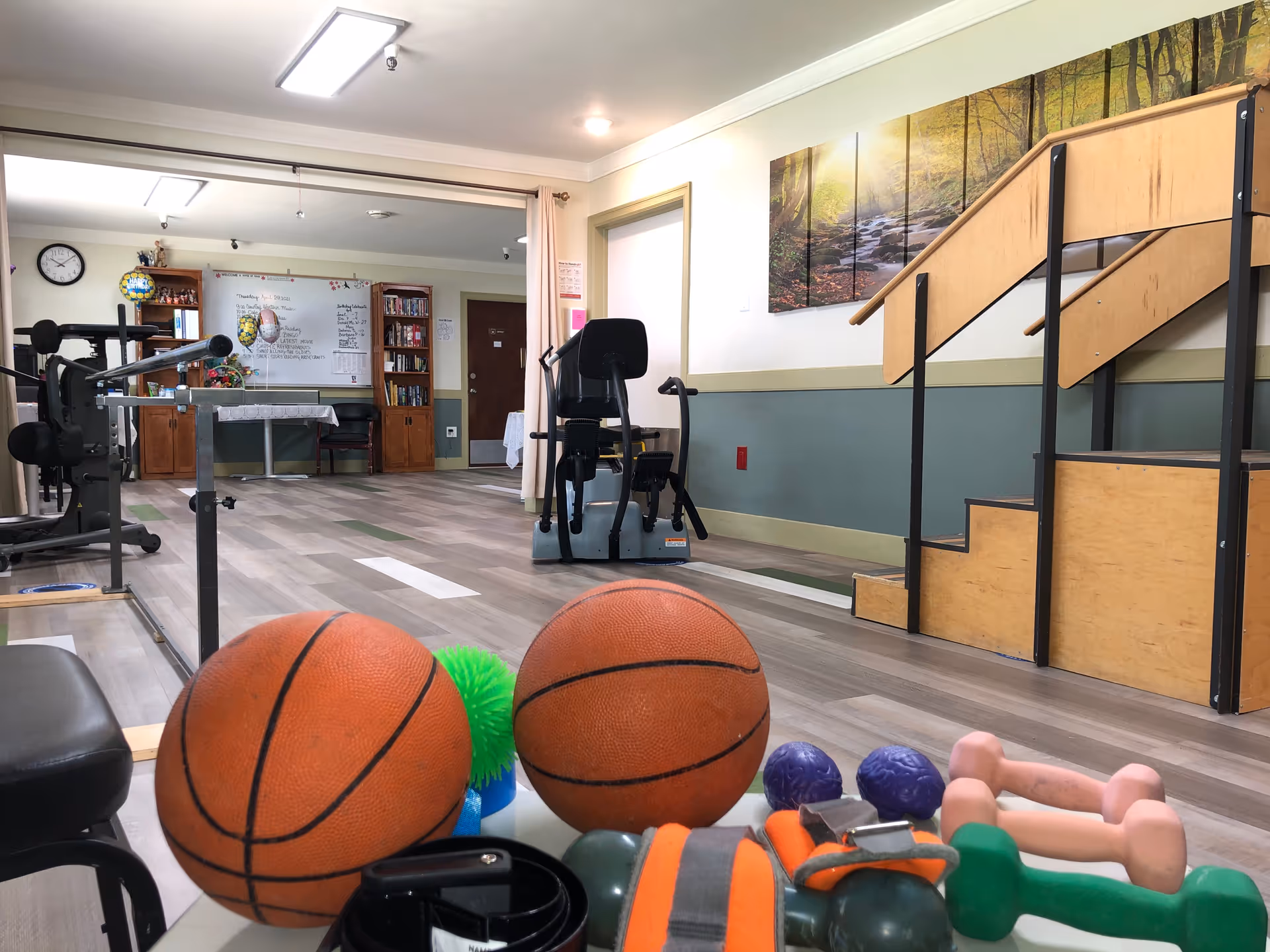 Indoor rehabilitation gym with workout equipment, stairs, and colorful balls in foreground.