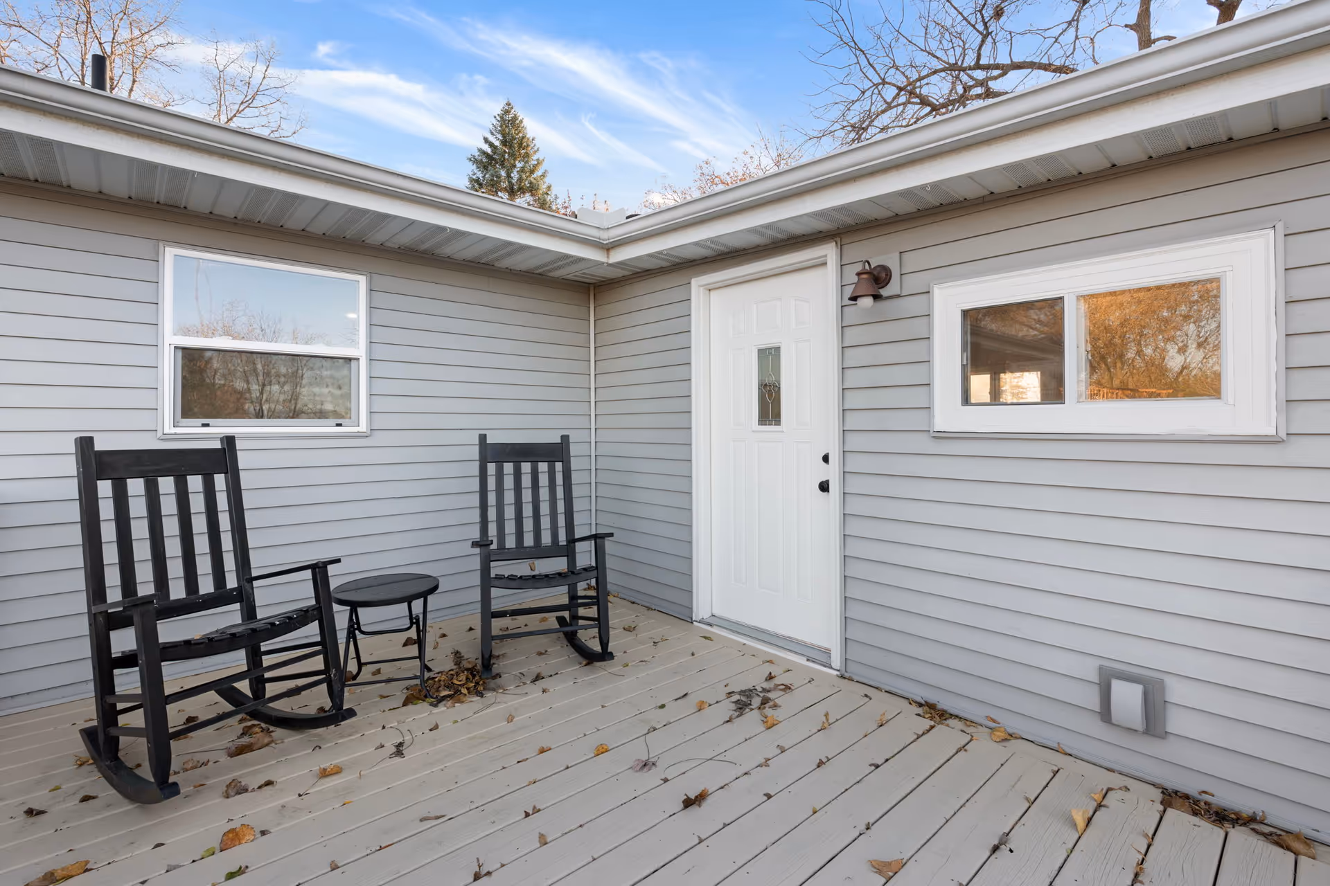 Two black rocking chairs and a small table on a wooden deck outside a gray building.