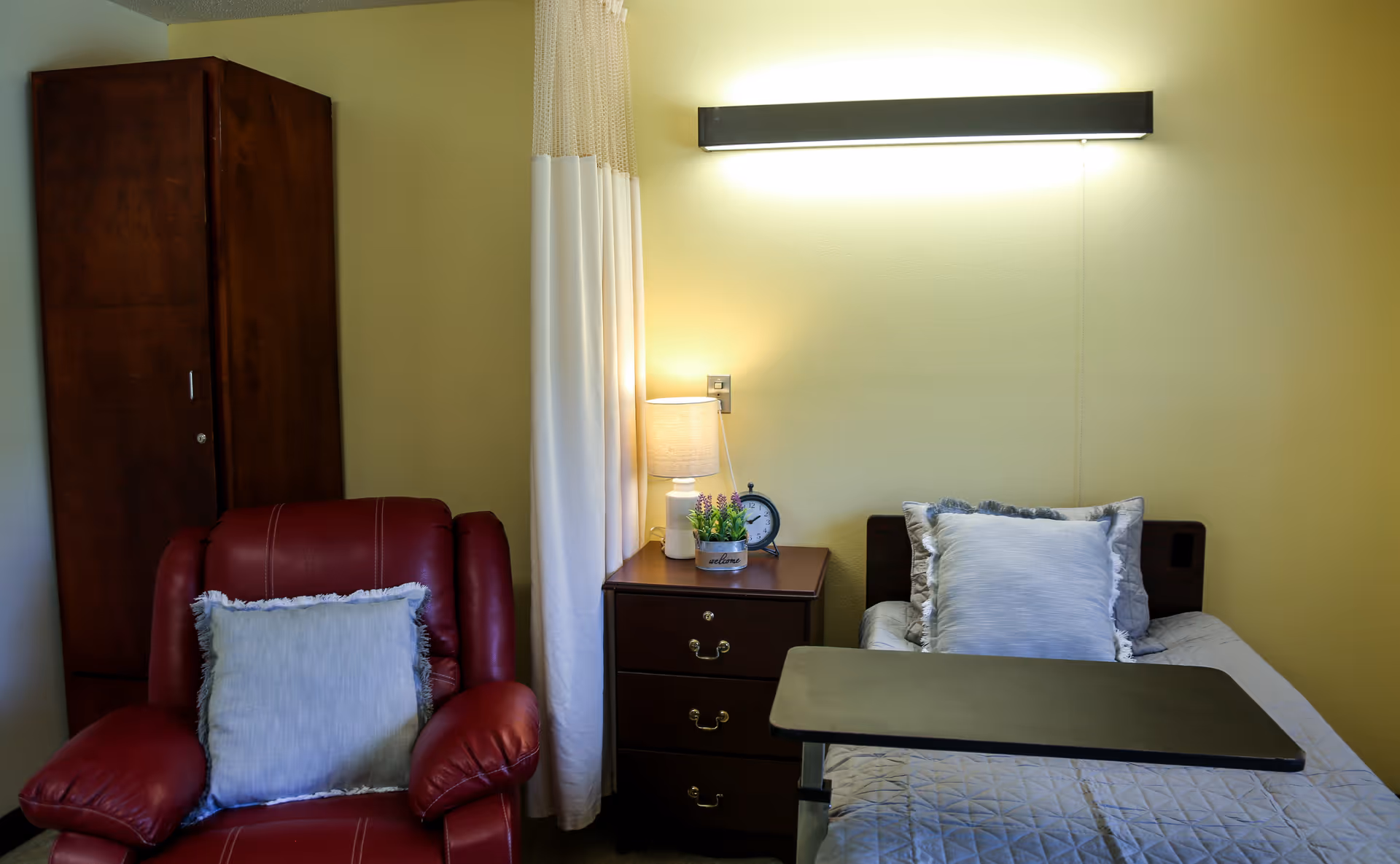 A cozy room with a single bed featuring pillows and a quilt, a bedside table with a lamp, a small plant, and an alarm clock. Next to the bed is a red armchair with a light-colored pillow. A wooden cabinet is visible in the corner, and a curtain hangs between the bed and the cabinet. The walls are painted light yellow.