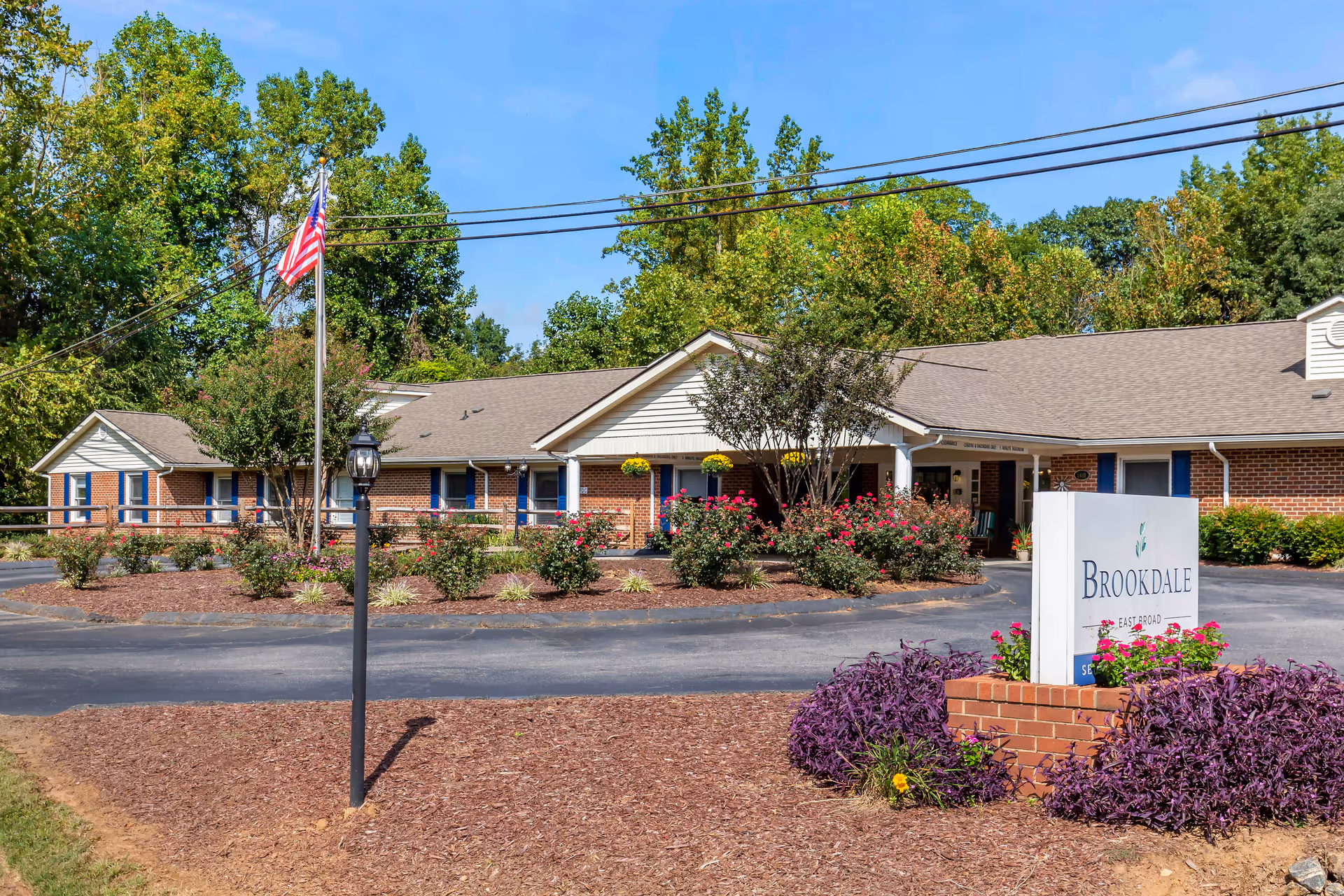 Exterior view of Brookdale East Broad senior living facility showing a single-story brick building with a pitched roof, surrounded by landscaped bushes and flowers. An American flag is flying on a flagpole near the entrance, and a sign with the facility's name is visible in the foreground.