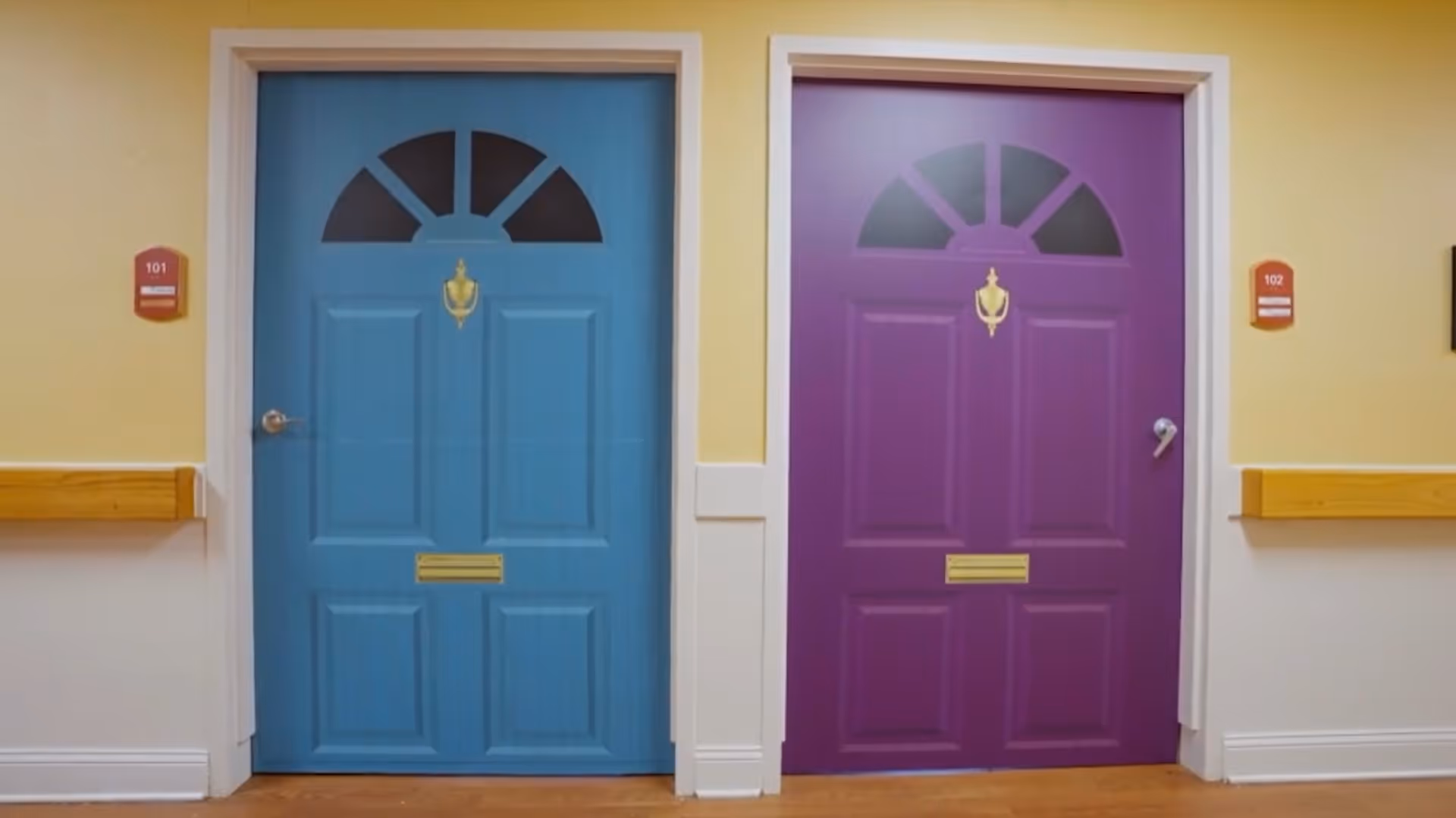 Two closed doors in a hallway, one painted blue and the other purple, each with a brass door knocker and mail slot. The blue door is labeled 101 and the purple door is labeled 102. The walls are painted yellow and white with wooden handrails on each side.