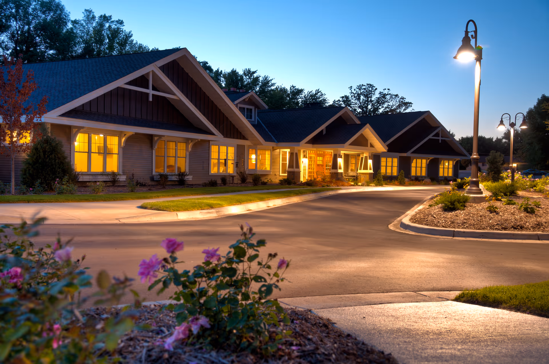 Exterior view of Benedictine Lvg Comm Winsted with lit windows and curved driveway at dusk.