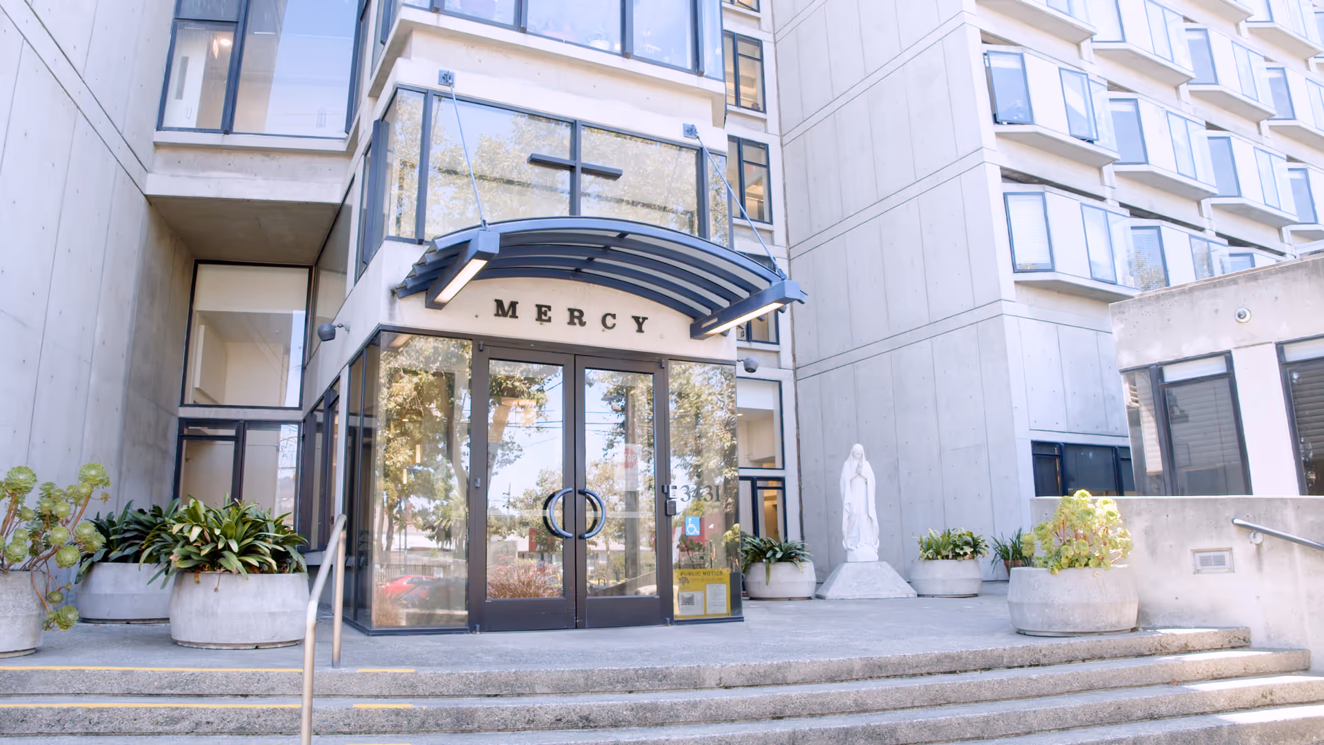 Front entrance of the Mercy retirement and care center with glass double doors under a curved awning, concrete steps, potted plants, and a statue.