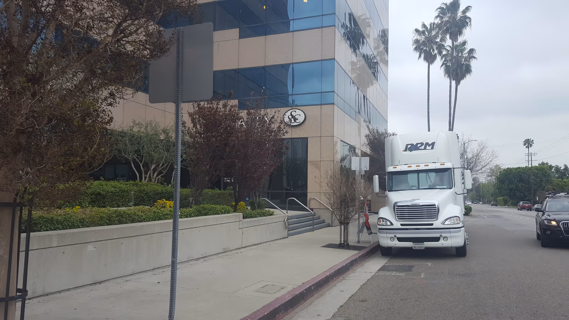 Modern glass-front building beside a sidewalk with a white semi-truck parked along the curb.