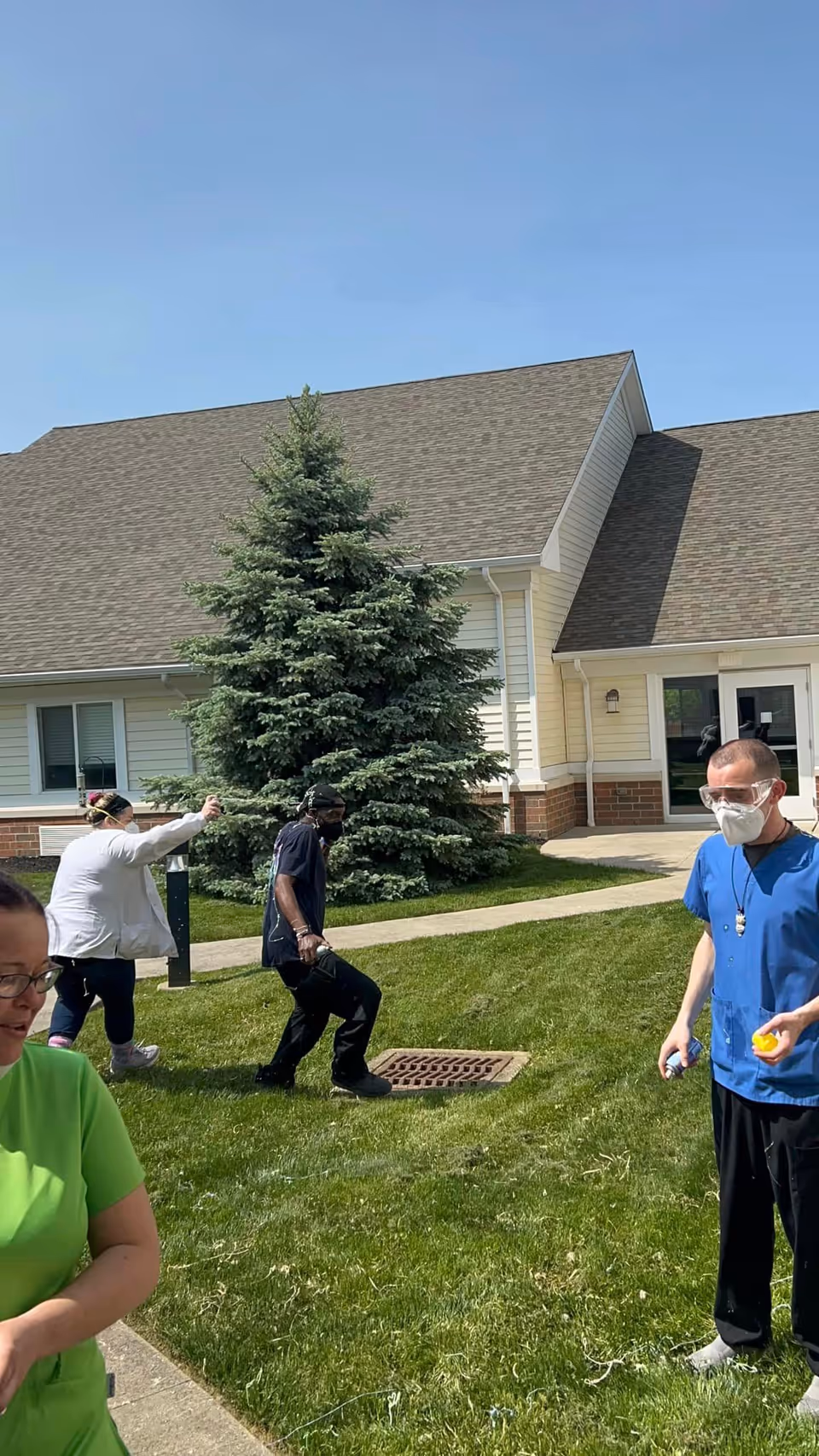Several people outside on a grassy area near a building with beige siding and a large evergreen tree. One person in blue scrubs and a face mask is holding small objects, while others are walking or standing on the grass and sidewalk under a clear blue sky.