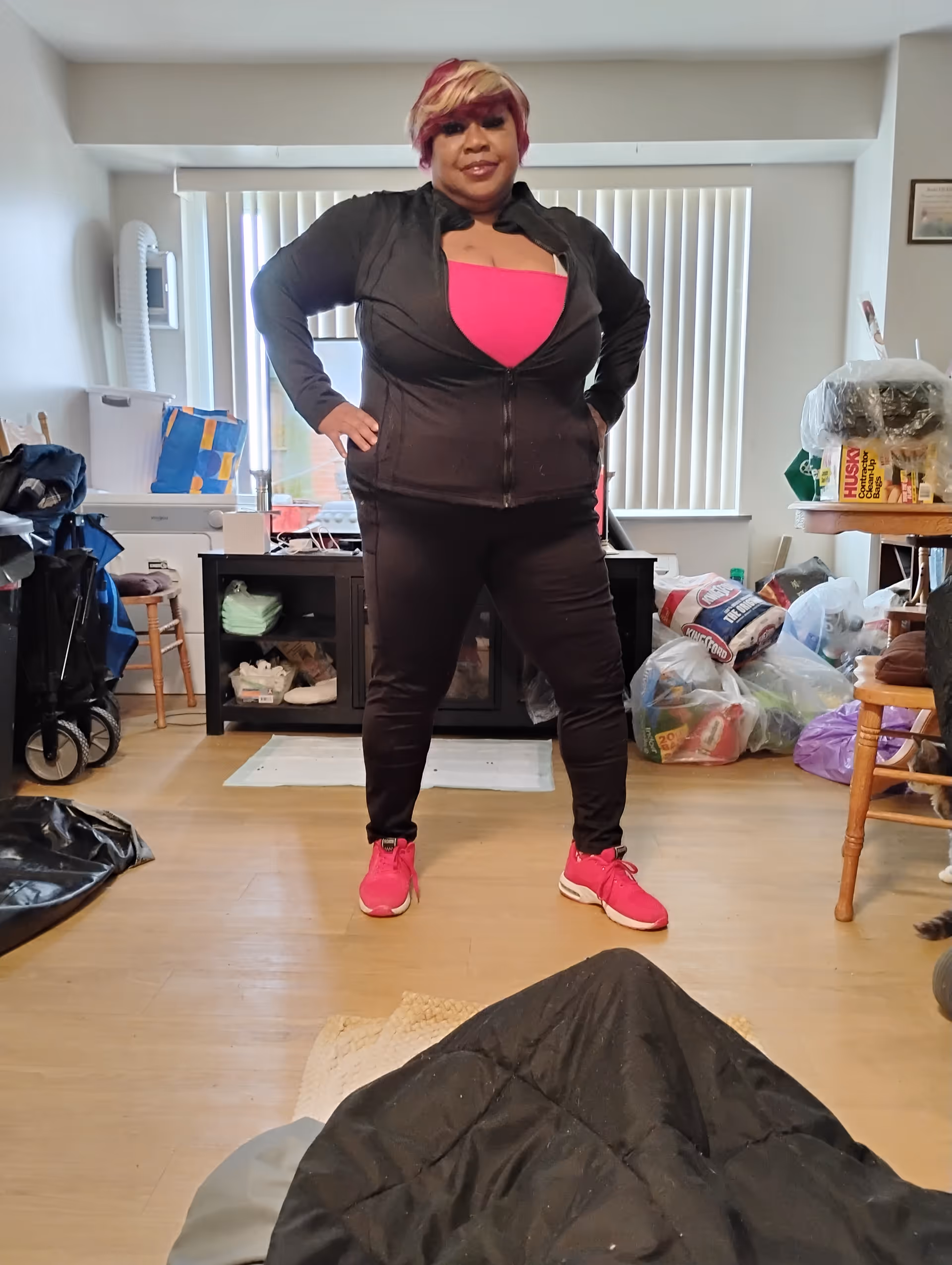 Person stands with hands on hips in a cluttered indoor living area.