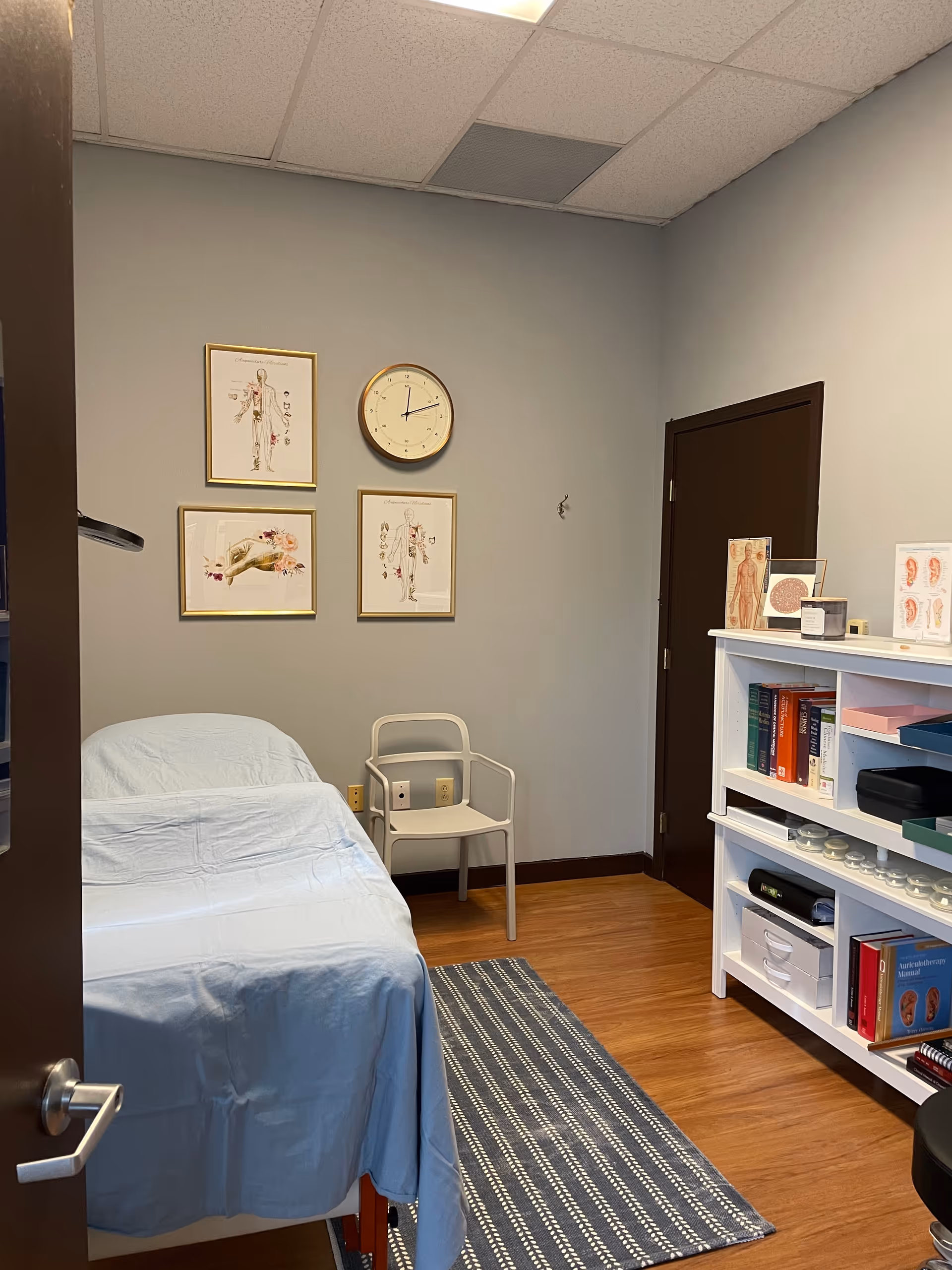 An interior exam room with a blue-covered bed, chair, bookshelf, and medical charts.