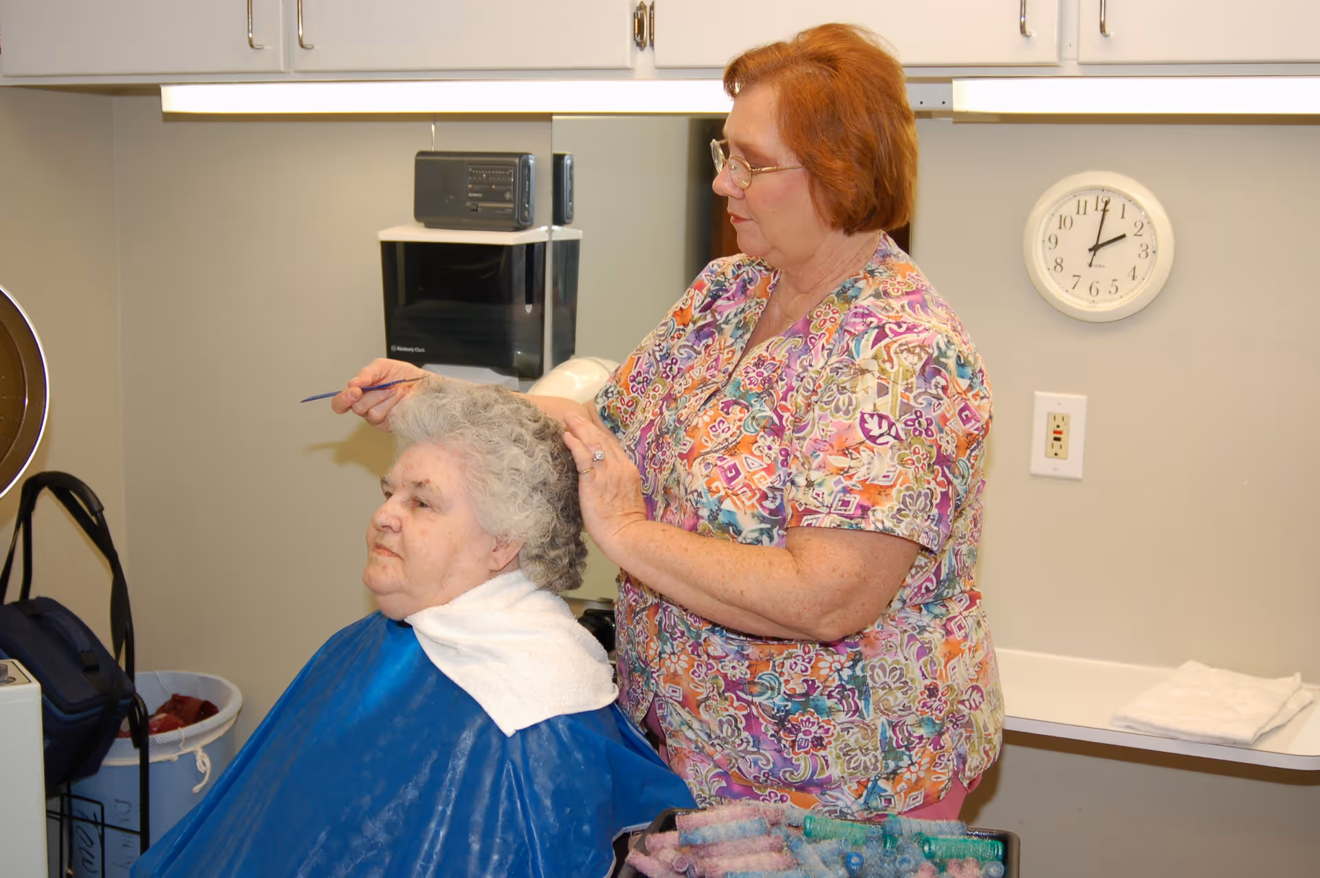 A senior woman with gray hair is seated and wearing a blue cape while another woman with red hair and glasses styles her hair using a comb in a room with white cabinets and a clock on the wall.