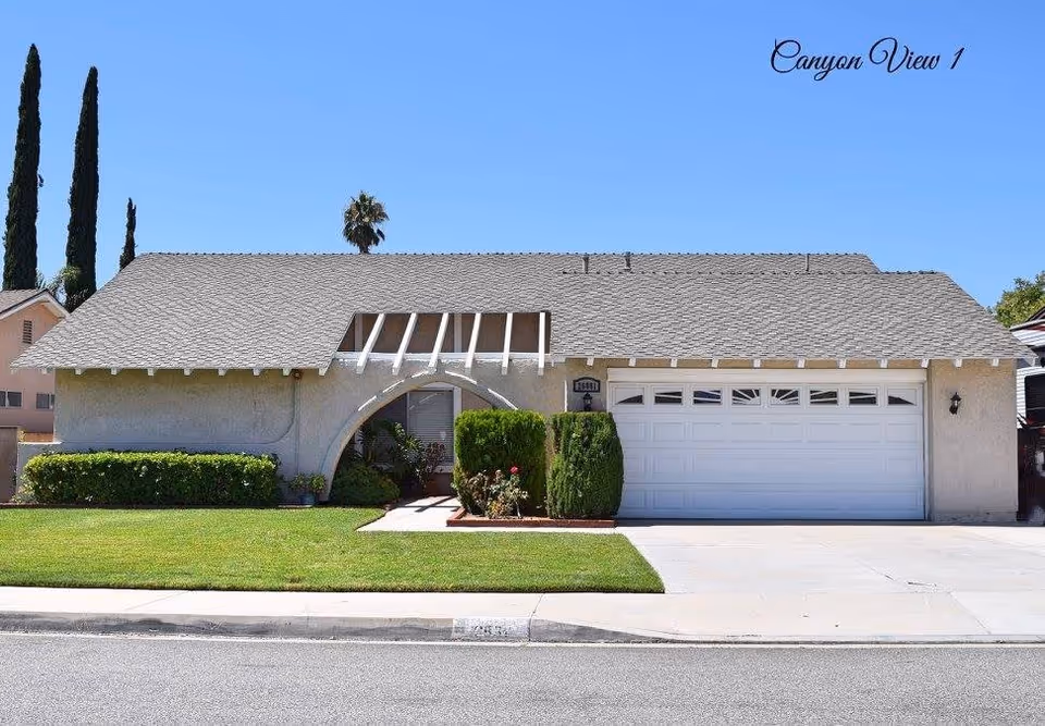 Single-story residential building with a gray shingled roof, white garage door, and an arched entrance surrounded by green bushes and a well-maintained lawn under a clear blue sky.