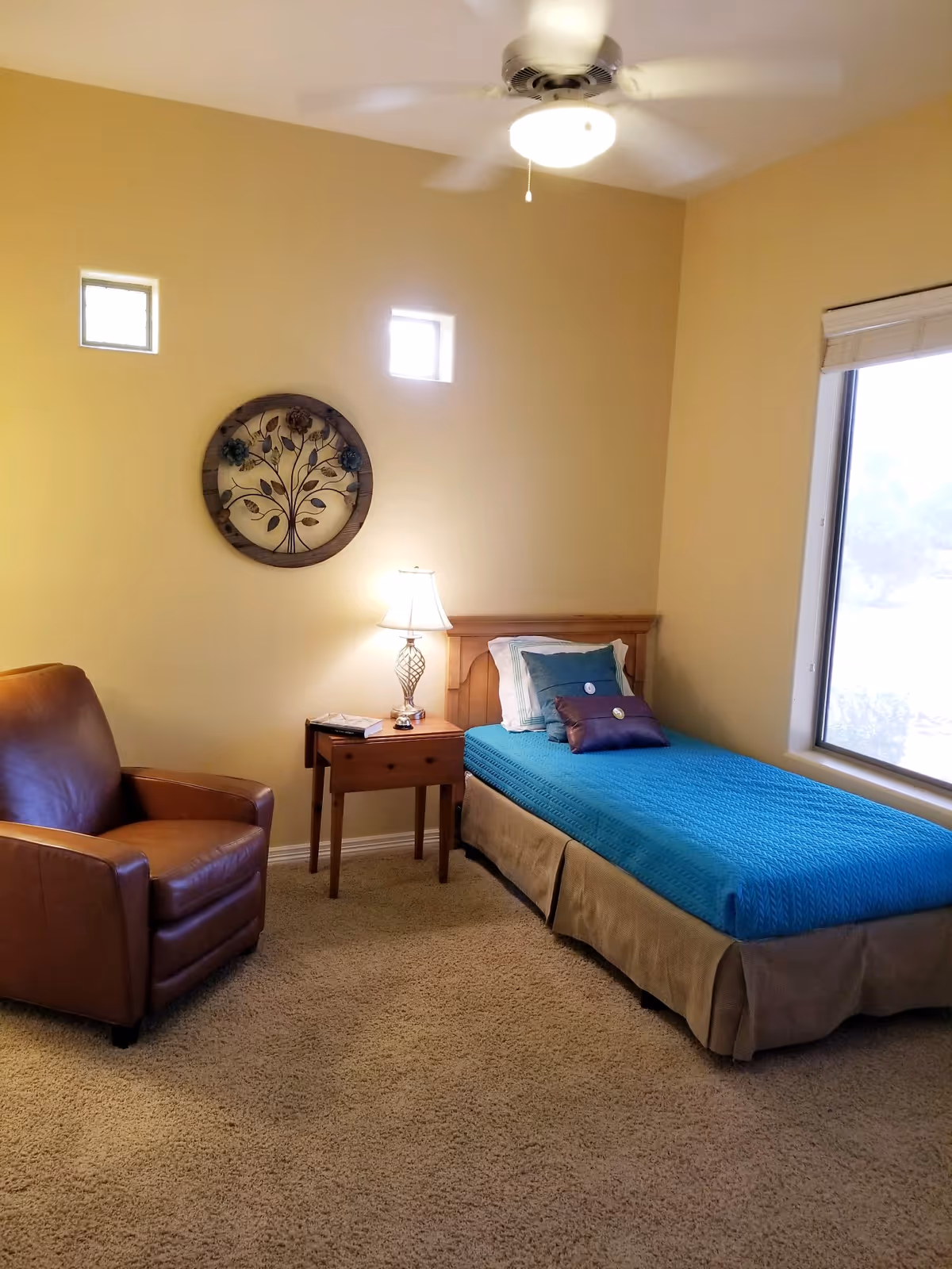 A cozy bedroom with a single bed covered in a blue quilt and decorative pillows, a wooden nightstand with a lamp and a book, a brown leather armchair, beige carpet, and a window with a view outside. The walls are painted light yellow and decorated with a round metal wall art piece featuring a tree design. A ceiling fan with a light is mounted on the ceiling.