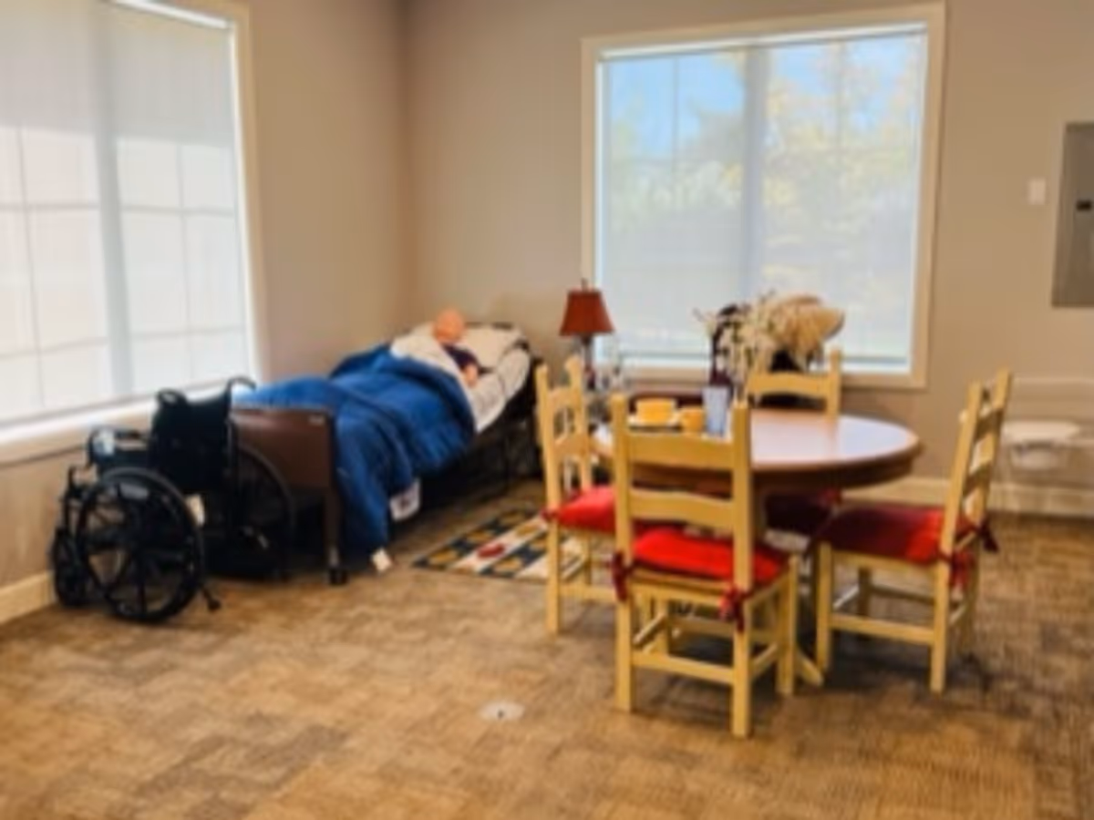 A resident rests in bed beside a wheelchair in a well-lit common interior room.