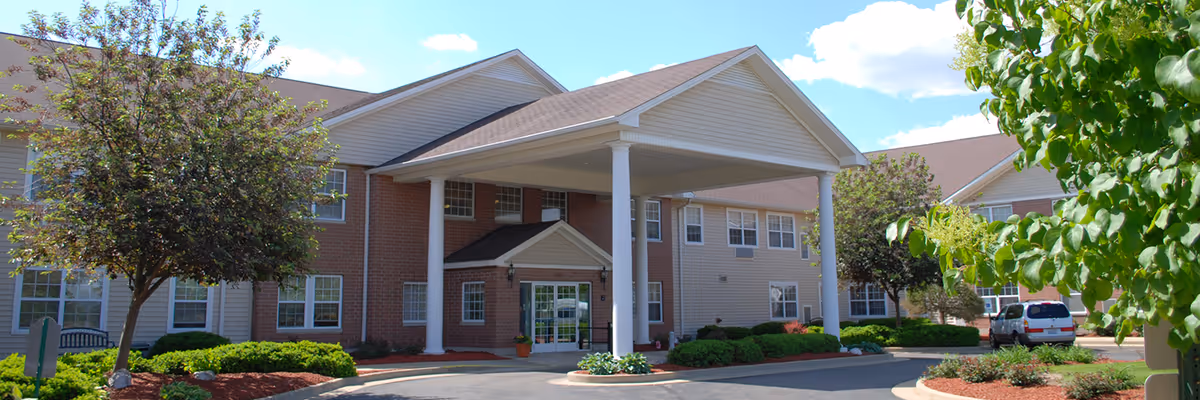 Front exterior view of a senior living facility building with a covered entrance supported by white columns, surrounded by landscaped greenery and trees under a blue sky with some clouds.