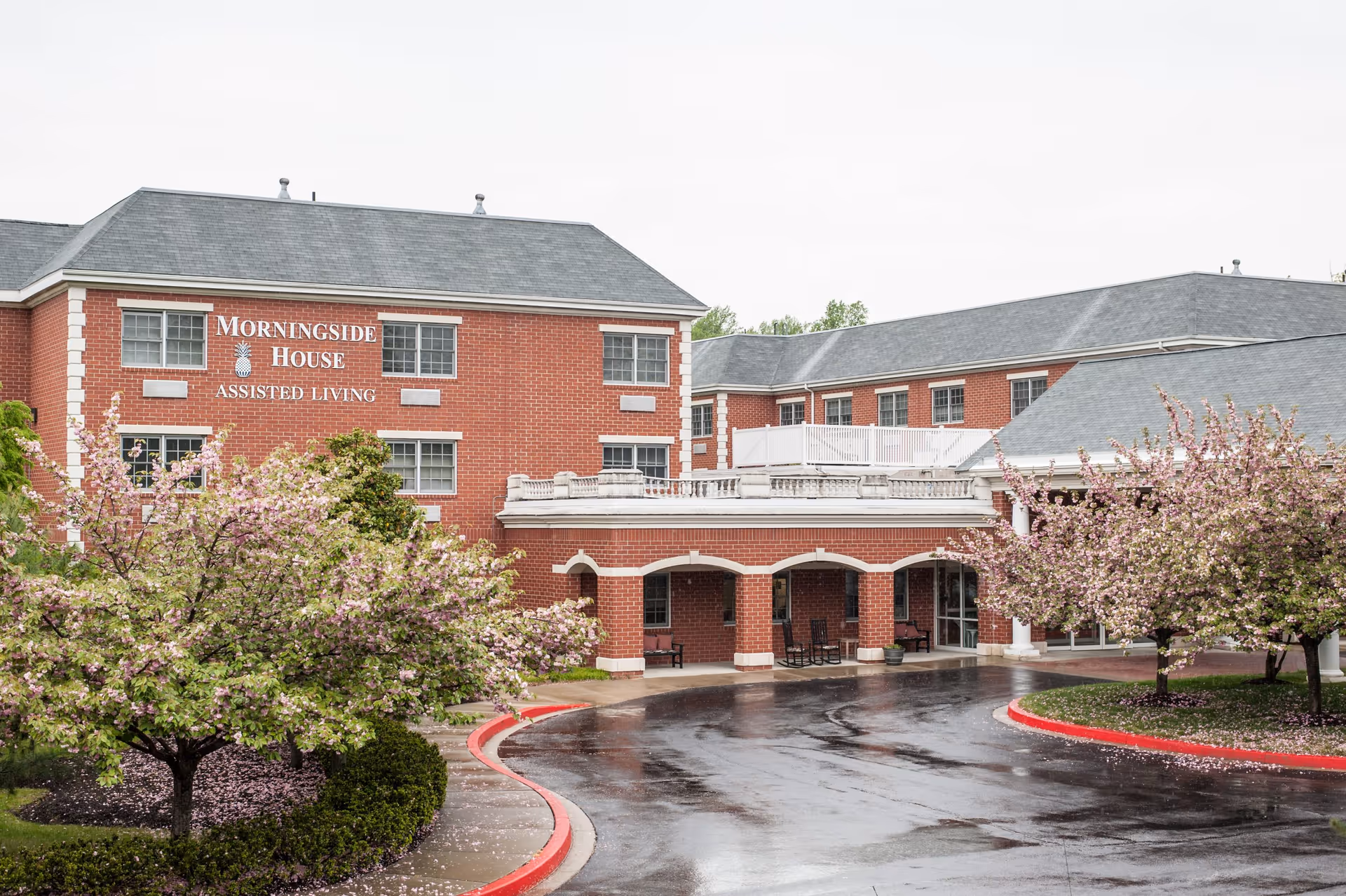 Front exterior of a red-brick assisted living building labeled "Morningside House" with flowering trees and a wet driveway.