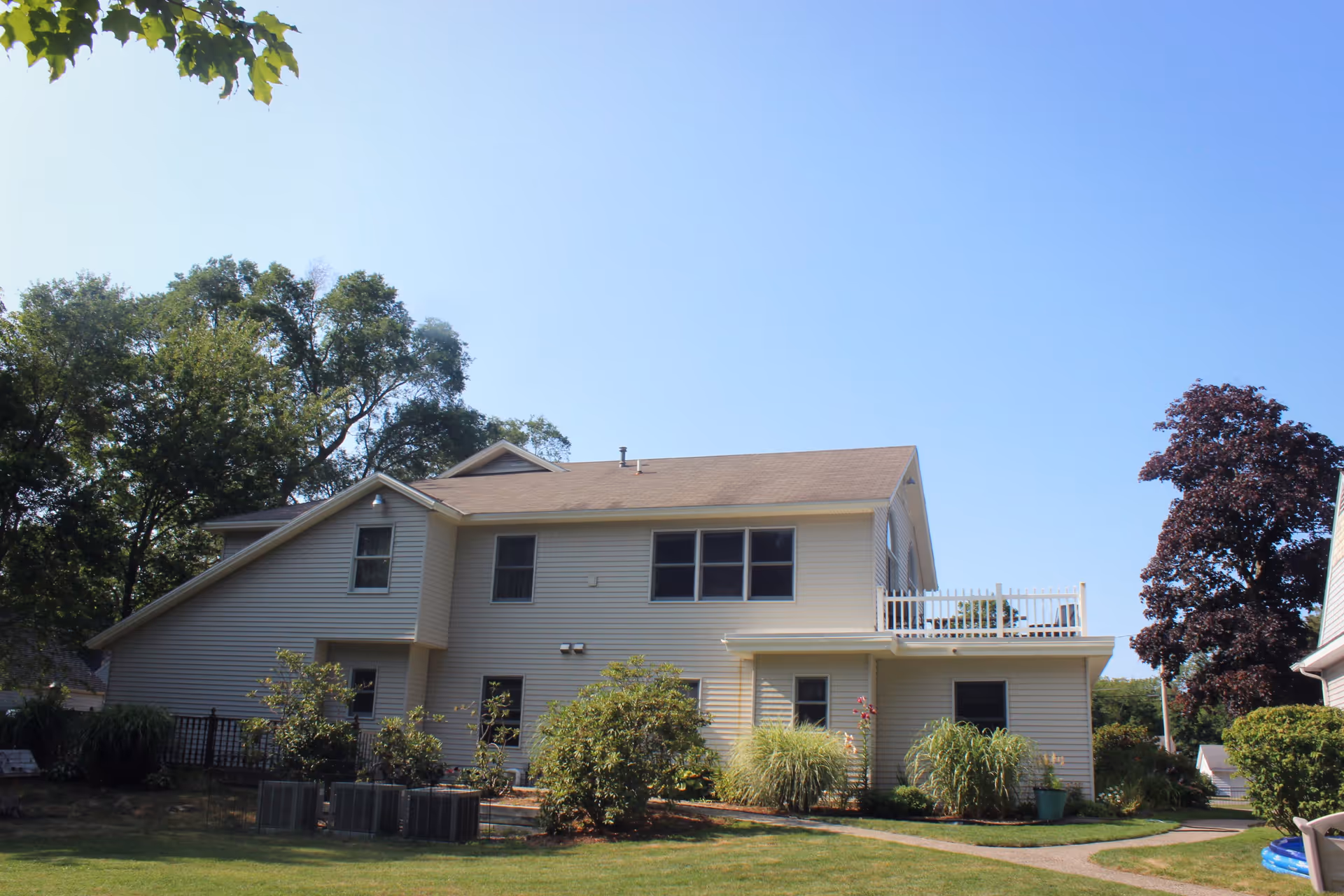 Exterior view of a two-story beige residential building with multiple windows, surrounded by green trees and shrubs under a clear blue sky.