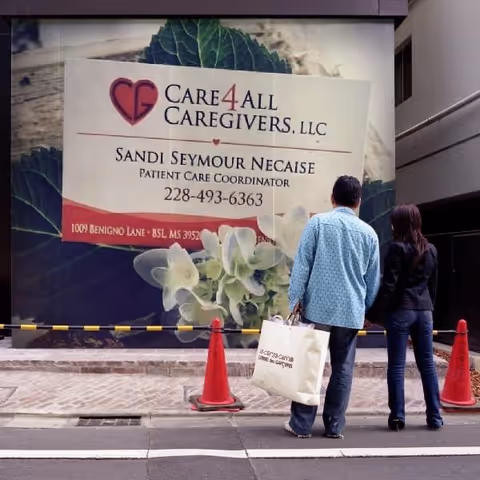 Two people stand outside a building with a large Care4All Caregivers sign.