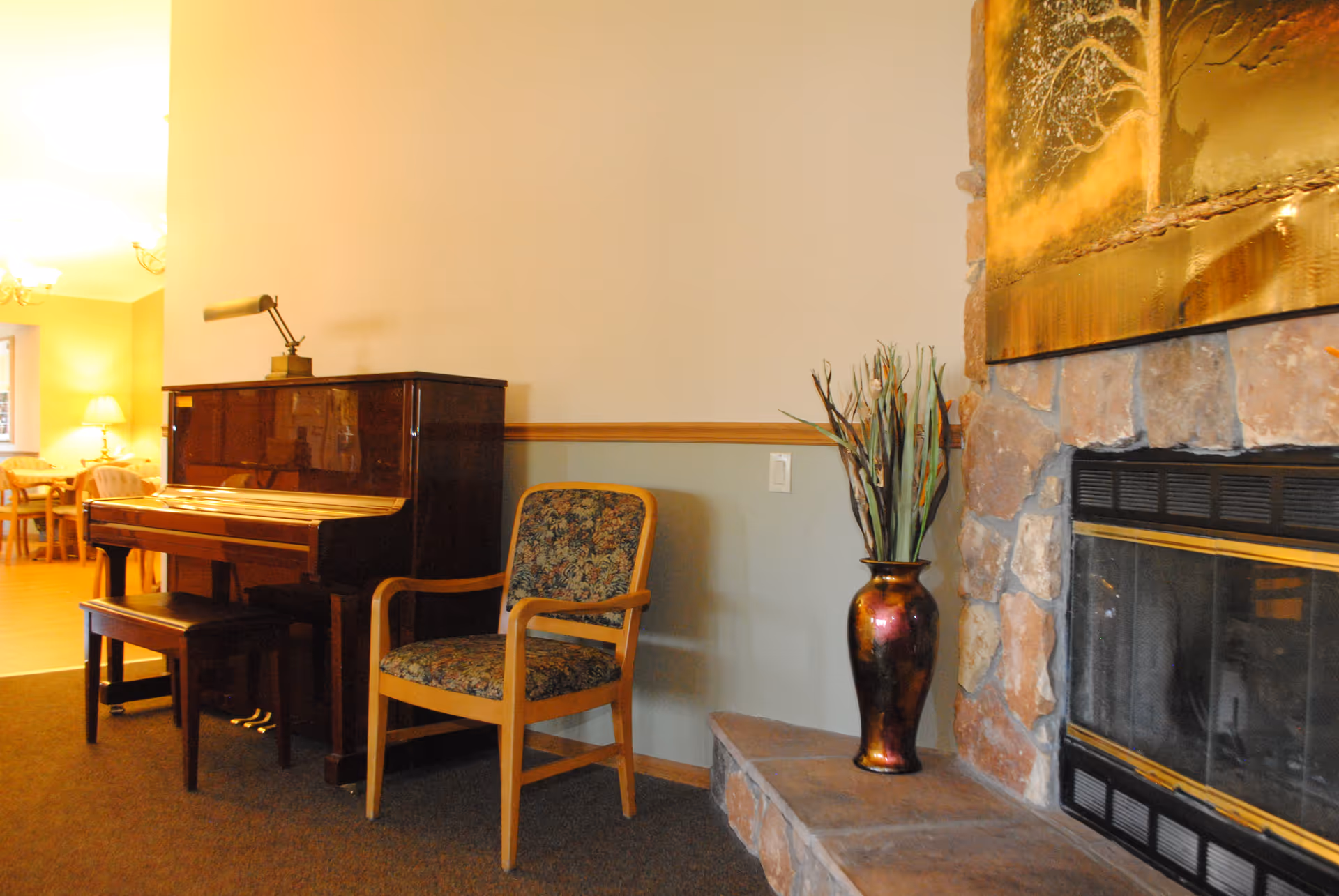 Interior common living area with an upright piano, a floral-upholstered chair, a decorative vase on a hearth, and a stone fireplace with dining area visible in the background.