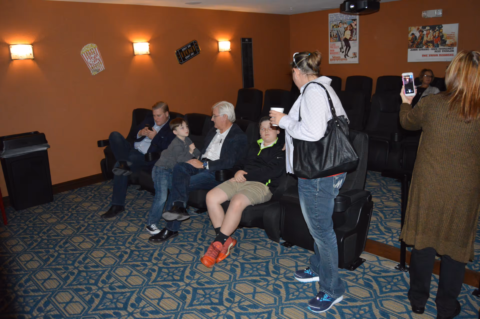 A group of people sitting and standing in a small theater room with orange walls and patterned blue carpet. There are movie posters on the wall and wall-mounted lights. Some people are seated in black theater chairs, while others are standing, one holding a cup and another taking a photo with a smartphone.