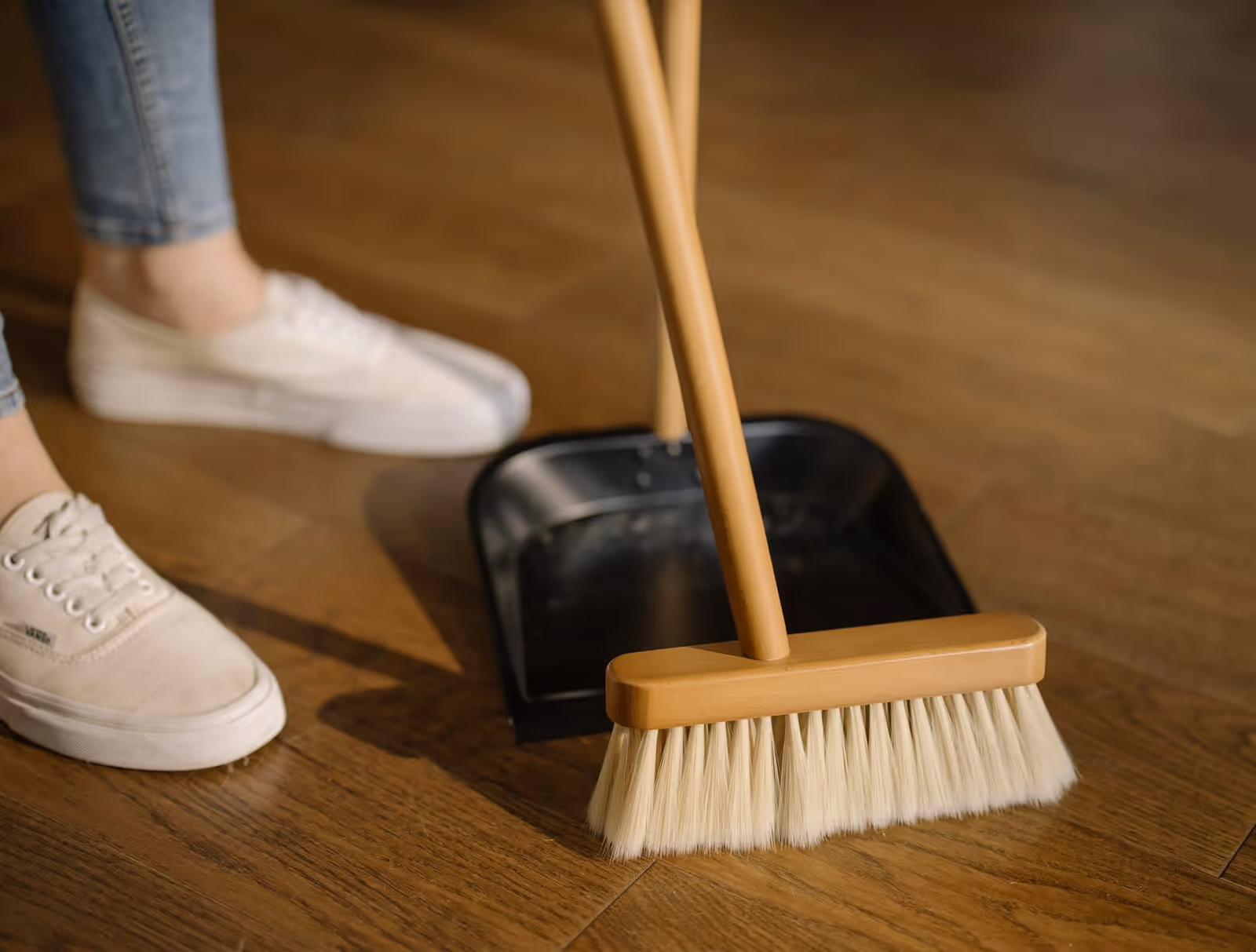 A person stands near a broom and dustpan on a wooden floor.