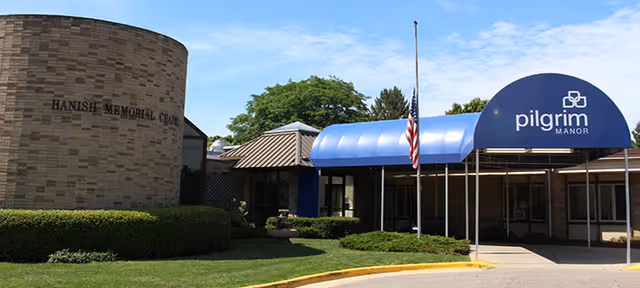 Exterior view of a senior living facility entrance with a blue awning labeled 'pilgrim MANOR' and a flagpole with an American flag. The building has a brick wall with the text 'HANISH MEMORIAL CHAPEL' and is surrounded by green bushes and a lawn under a partly cloudy sky.