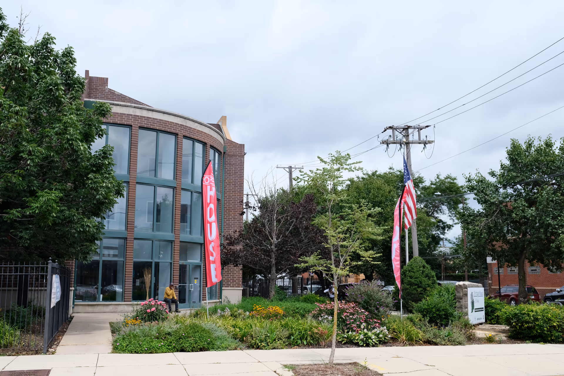 Exterior of a brick building with large curved glass windows, a landscaped front garden, and flags including an American flag.
