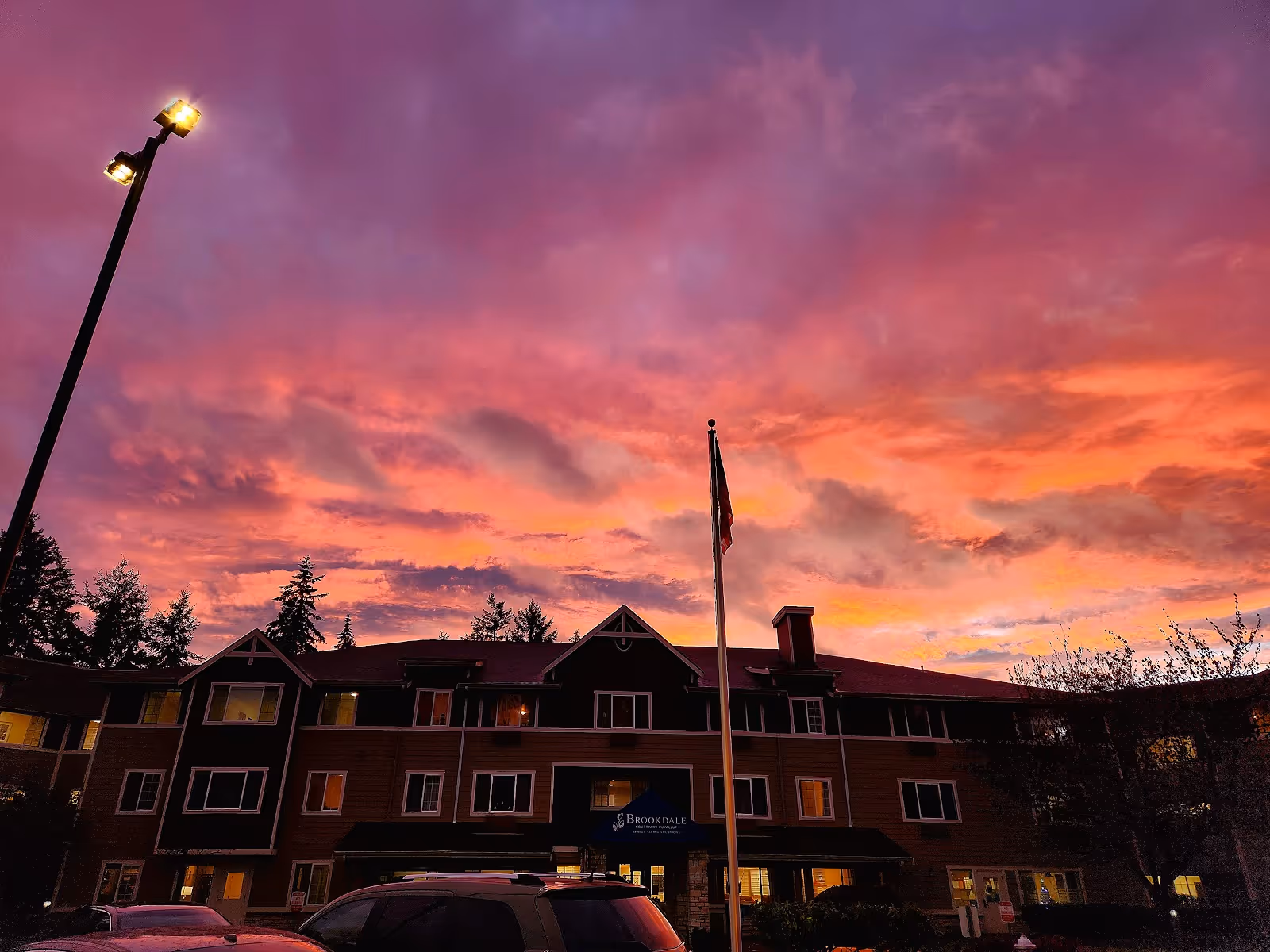 Sunset sky above Brookdale Courtyard Puyallup exterior with flagpole and parked cars.