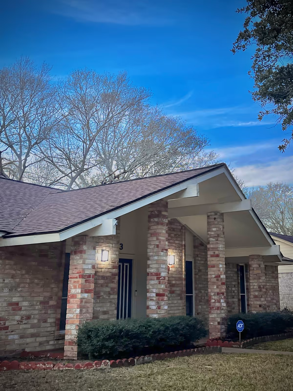 Entrance of Carnation Oaks, brick exterior with columns and shrubs under a blue sky.