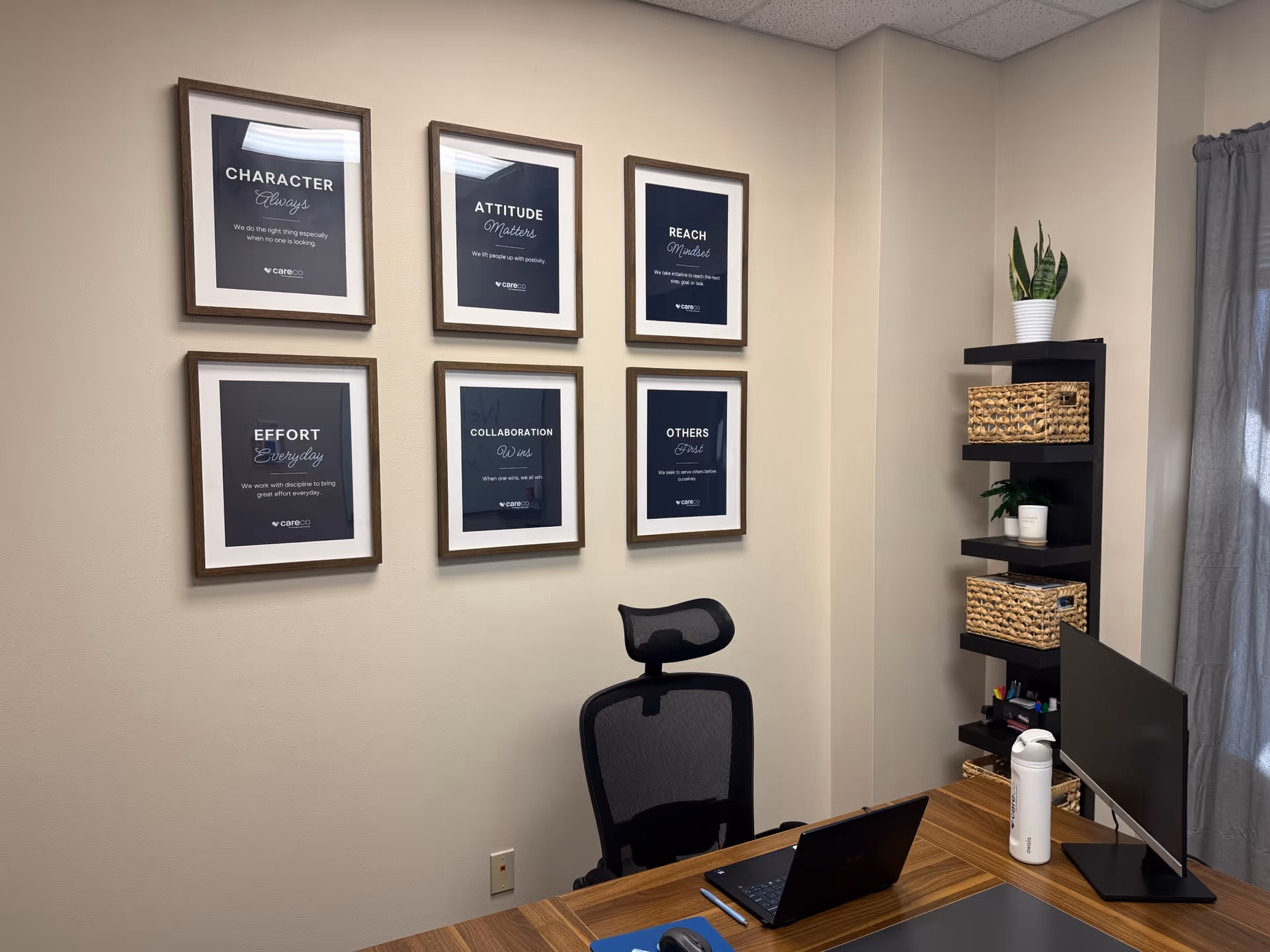 Office desk area with framed motivational posters, computer monitor, laptop, and shelving unit.