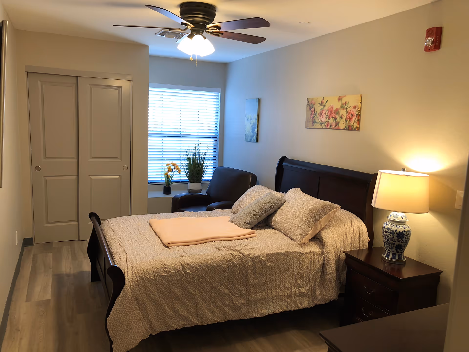 A cozy bedroom with a wooden bed frame, neatly made bed with patterned bedding and pillows, a folded blanket on top, a wooden nightstand with a blue and white ceramic lamp, a ceiling fan with lights, a window with blinds, two floral paintings on the walls, a dark armchair near the window, and a closet with sliding doors.