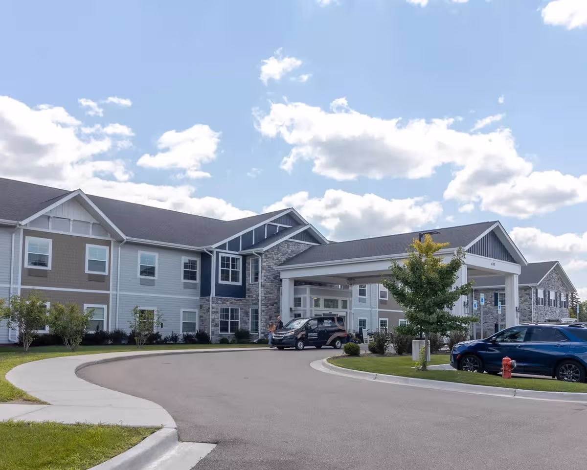 Modern senior living facility exterior with covered entrance, parked cars, and landscaped grounds.