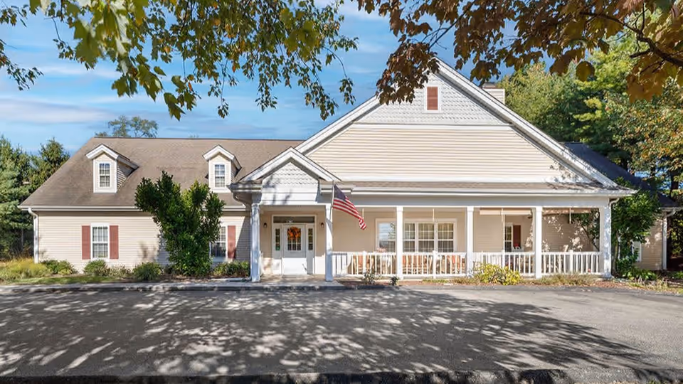 Front exterior view of a single-story building with beige siding, white trim, and a covered porch with white railings. An American flag is mounted near the entrance. Trees with green and brown leaves frame the top of the image, and there is a paved driveway in front of the building.