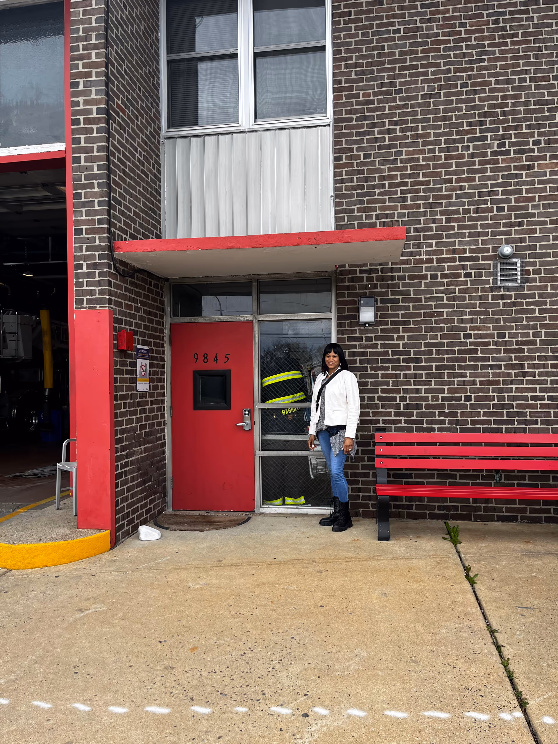 A woman stands outside a brick building entrance next to a red bench.