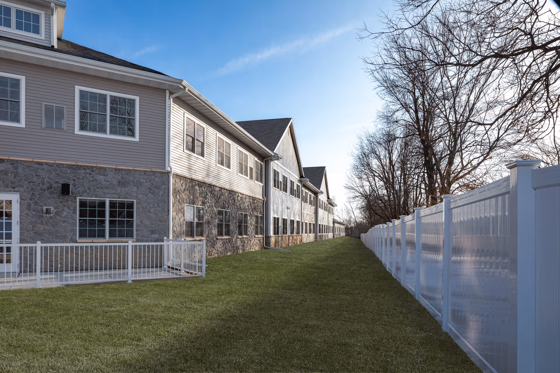 Side exterior of a two-story assisted living building with stone siding, a white fence, and a grassy yard under a clear blue sky.