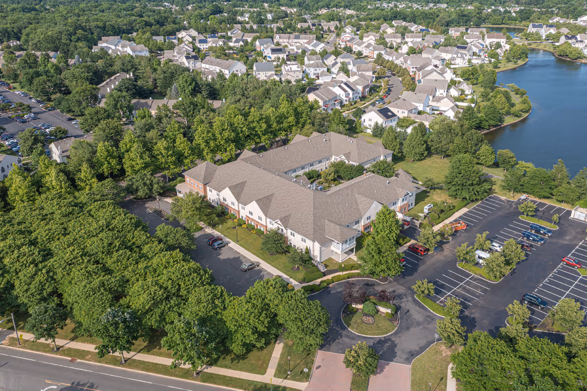 Aerial view of a senior living facility named Rose Hill surrounded by trees, parking lots, residential houses, and a nearby lake. The building is large with a complex roof structure and is situated in a green, suburban area.