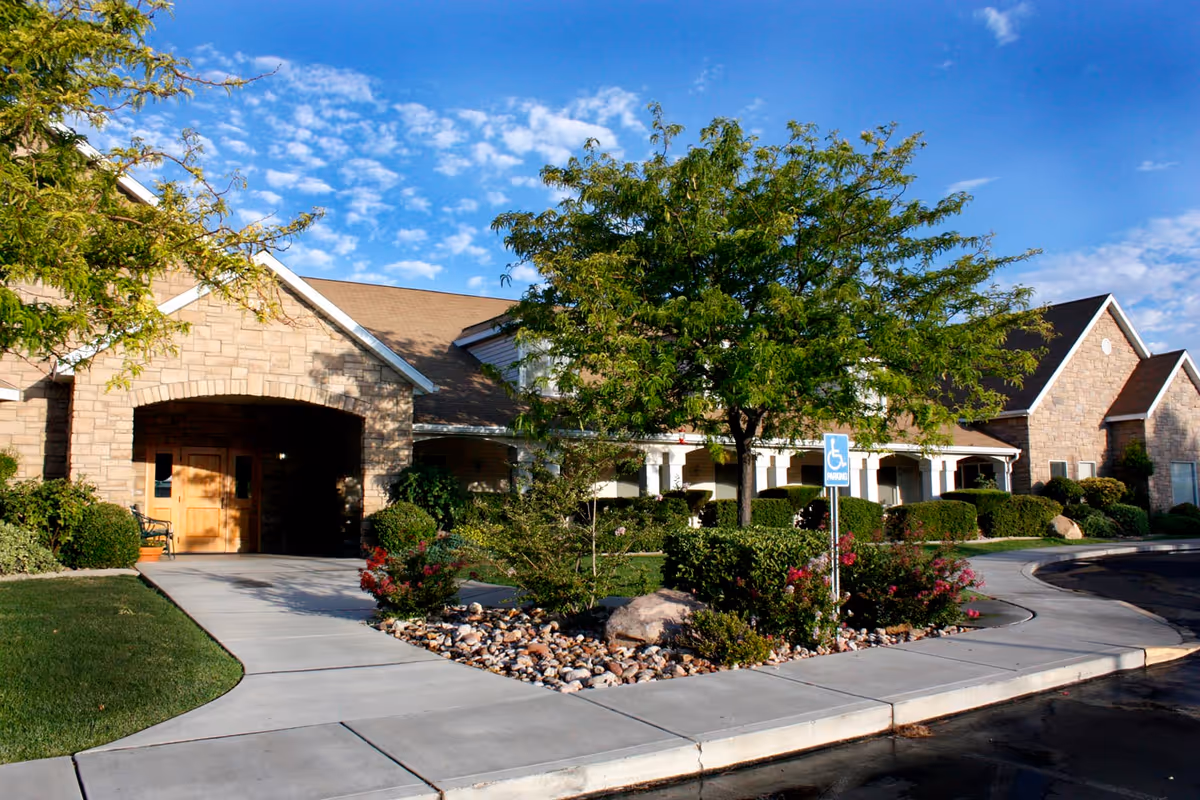 Front entrance of an assisted living building with a covered driveway, landscaped beds, a tree, and a handicap parking sign.