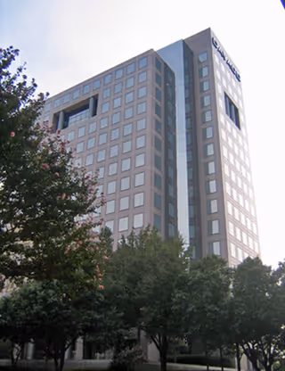 Tall modern office building with many windows, partially obscured by trees.