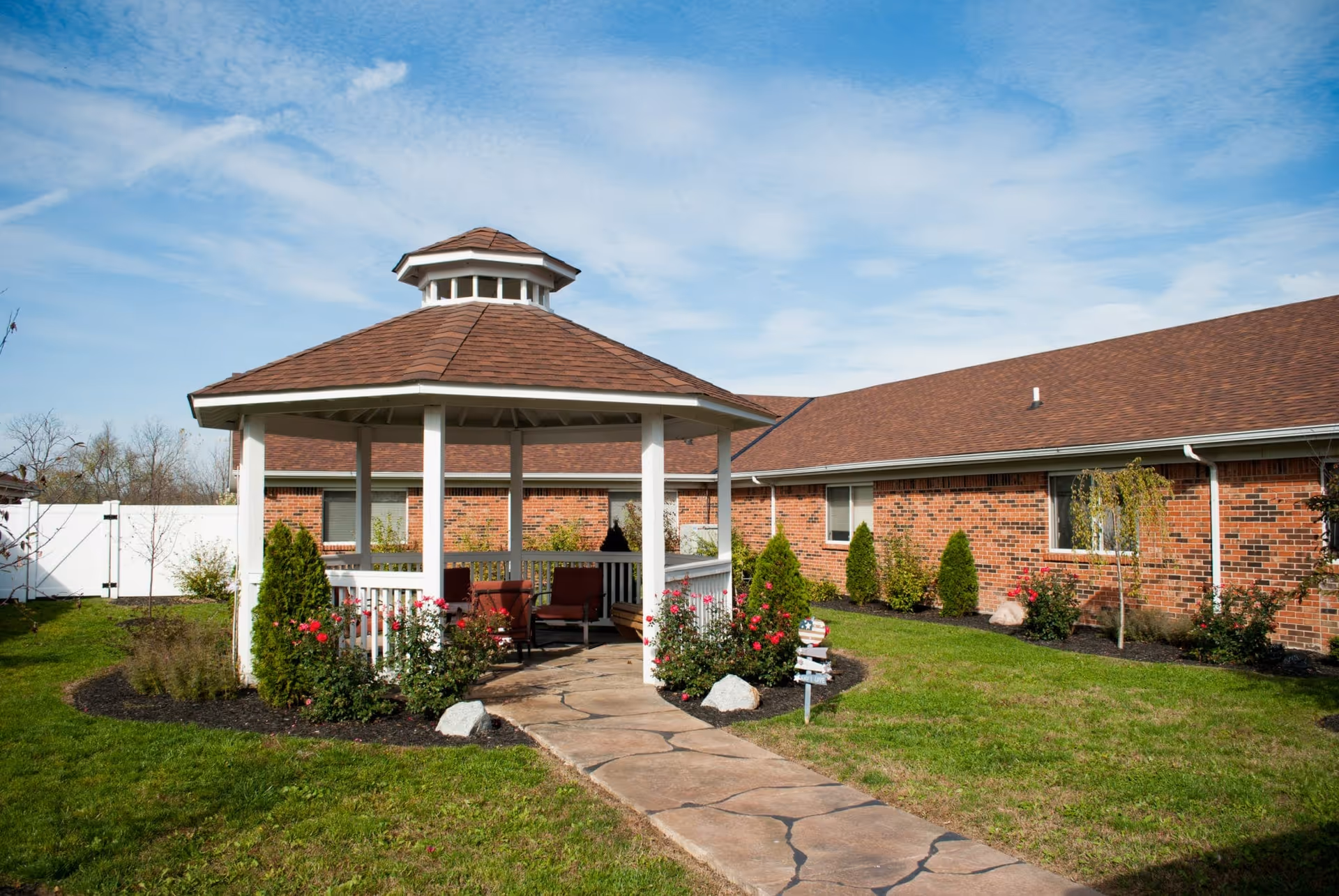 A white gazebo with seating stands in a landscaped courtyard in front of a single-story brick rehabilitation center under a blue sky.