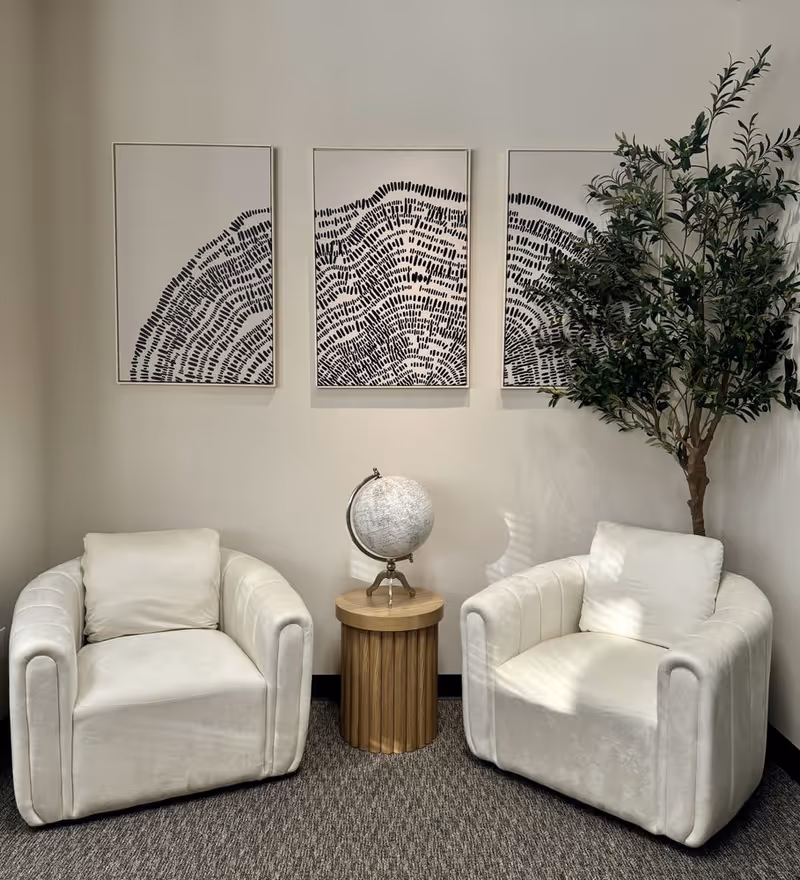 Two white armchairs flank a round wooden table with a globe, under three-panel wall art.