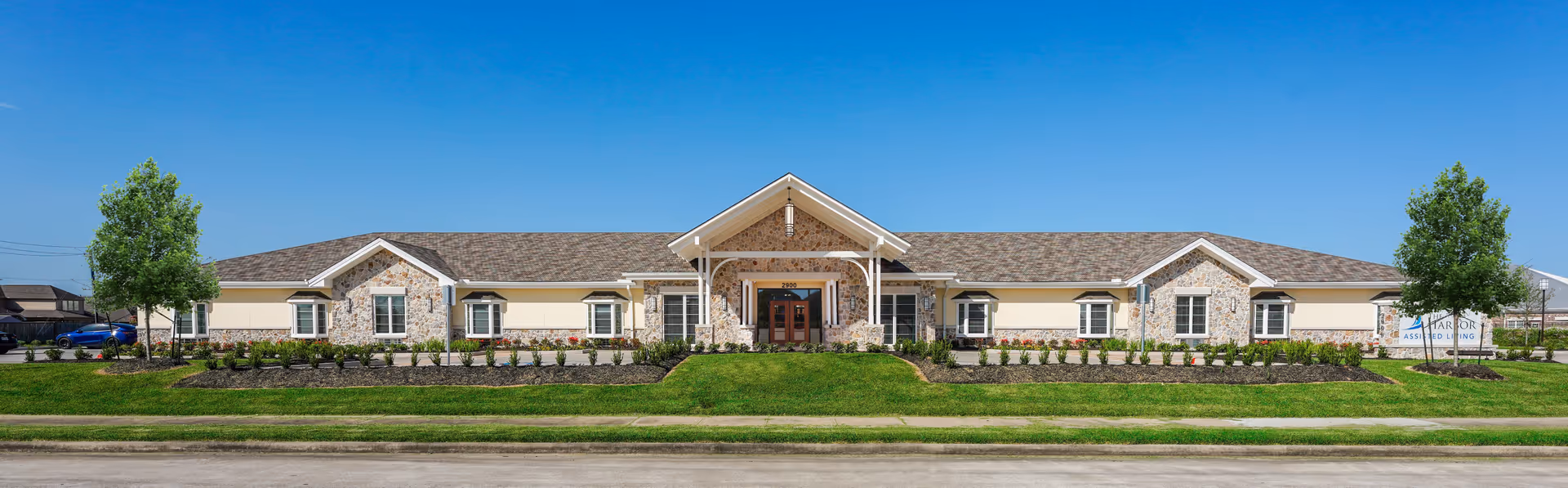 Front exterior of a single-story assisted living building with stone and siding facade, a central covered entrance and landscaped lawn under a clear blue sky.
