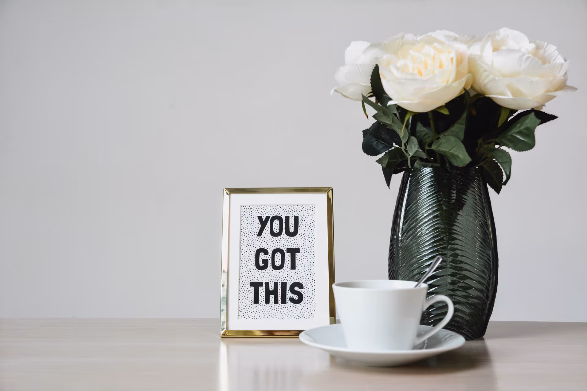 A gold-framed card beside a white teacup and a vase of white roses on a tabletop.