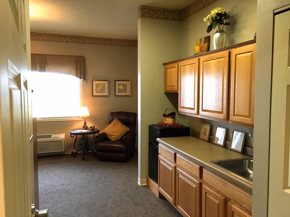 Interior view of a senior living facility room showing a small kitchenette area with wooden cabinets, a countertop, and a sink on the right side. In the background, there is a cozy sitting area with a brown recliner chair, a small round table with a lamp, and two framed pictures on the wall near a window with a valance.