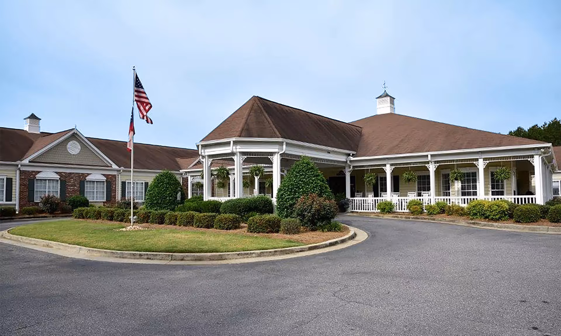 Exterior view of Viva Senior Living at Albany, showing a single-story building with a covered entrance, white columns, and a landscaped circular driveway with bushes and an American flag on a flagpole.