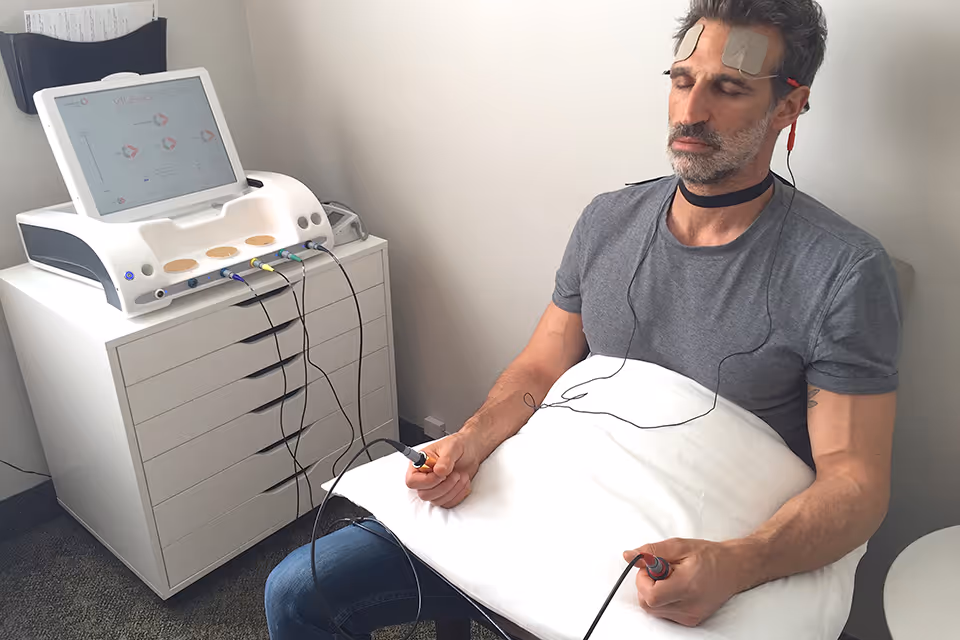 Man sits with pillow and EEG electrodes connected to a medical device in a clinical room.