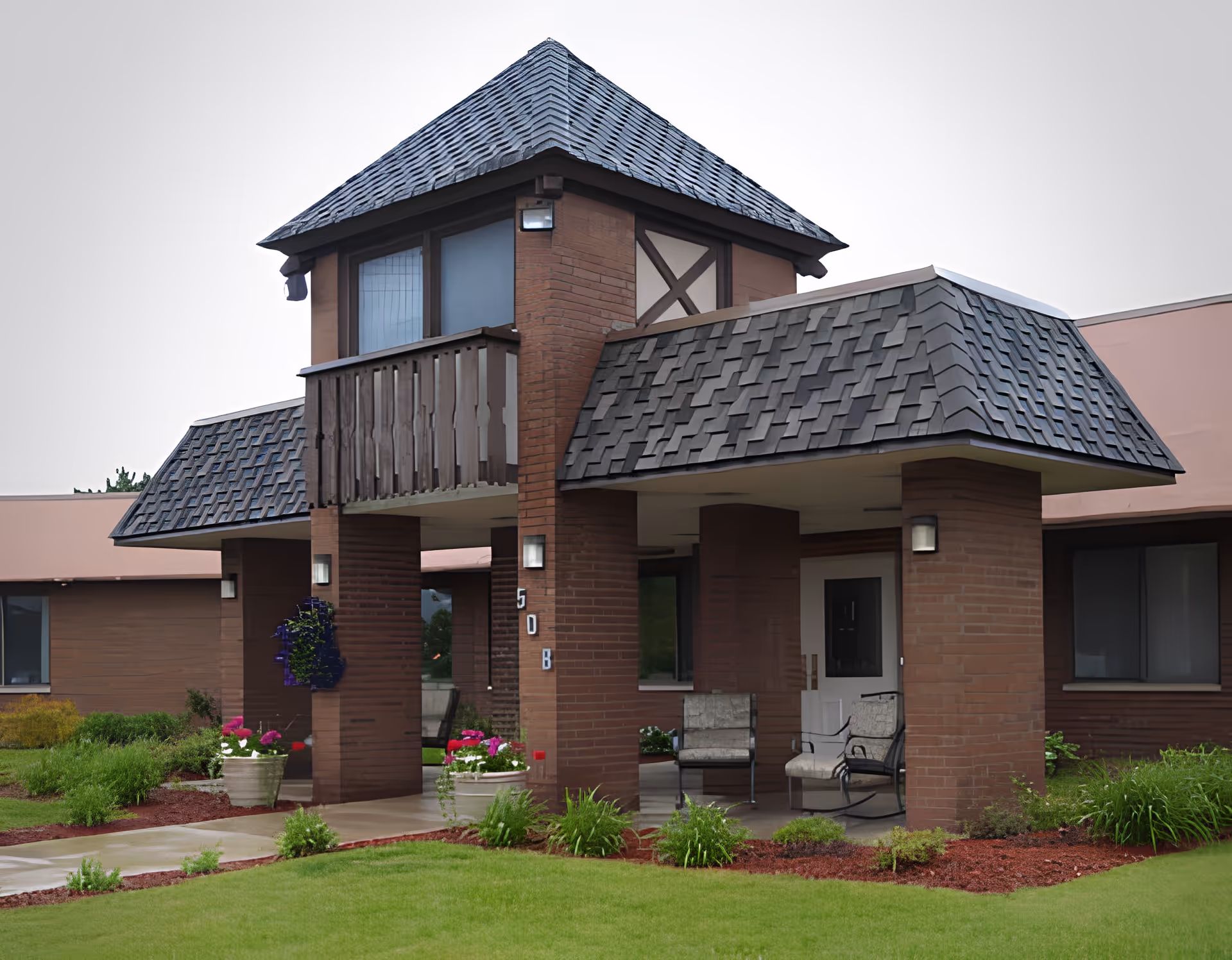 Exterior view of a building with a covered entrance supported by brick pillars, featuring a small balcony above. The building has a shingled roof and is surrounded by green grass and landscaped plants with flowers. There are chairs placed under the covered entrance area.