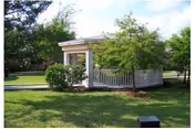 A small white building with a porch sits on a green lawn surrounded by trees.