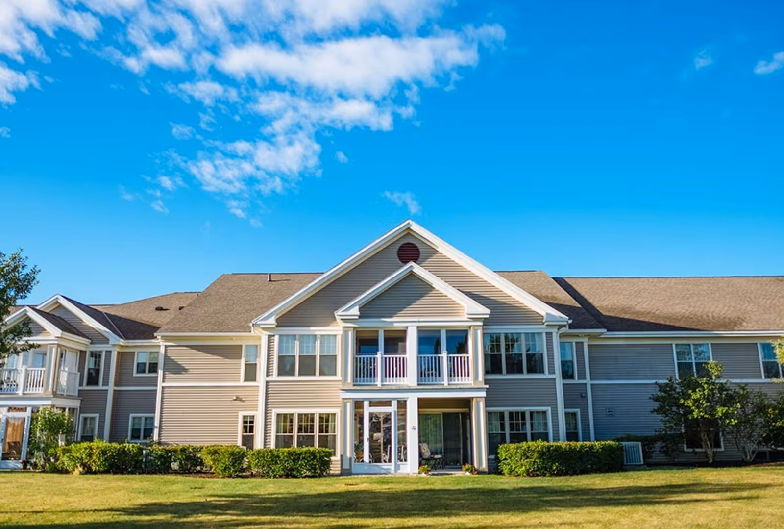 Front exterior of a two-story beige senior living building with balconies and a grassy lawn under a blue sky.
