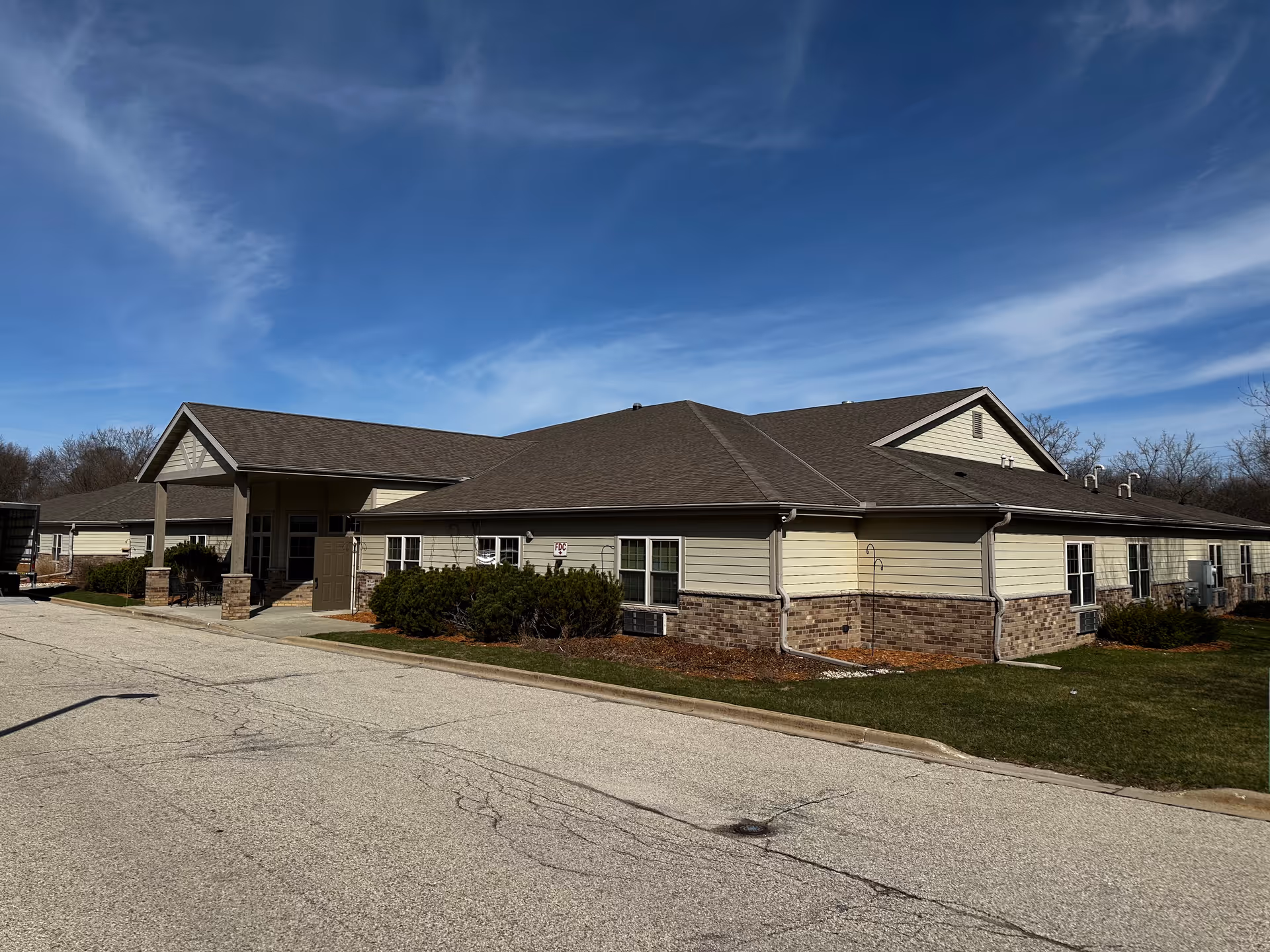 Exterior view of a single-story senior living facility building with beige siding and brick accents under a clear blue sky. The building has a covered entrance supported by columns and is surrounded by a paved driveway and some landscaping with bushes and grass.