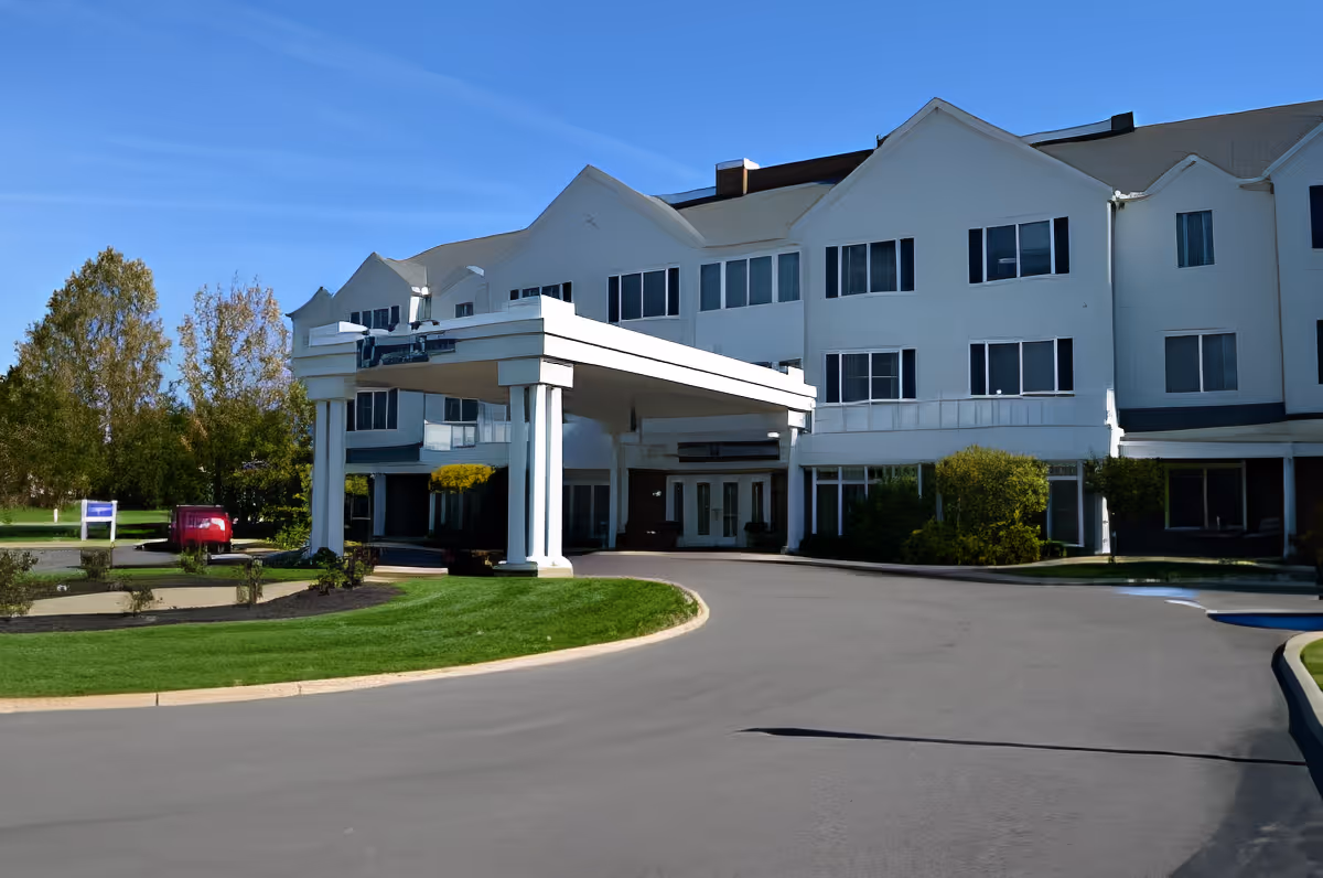 Front entrance of a white multi-story assisted living building with a covered porte-cochère and curved driveway.