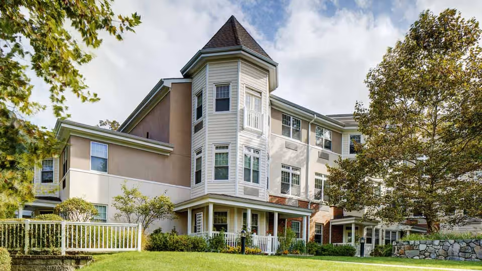 Multi-story senior living building with beige siding, front porch, and surrounding green lawn.
