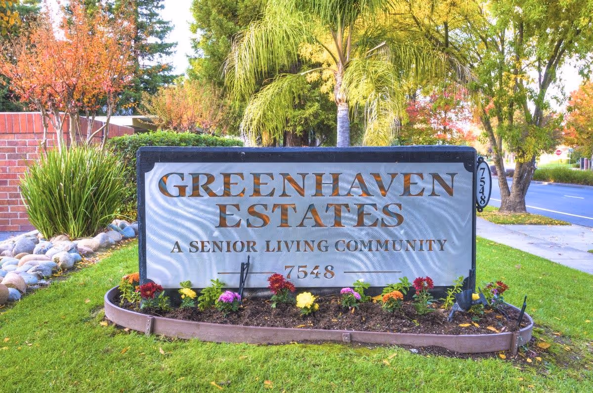 A large sign reads Greenhaven Estates, a Senior Living Community, surrounded by colorful flowers.