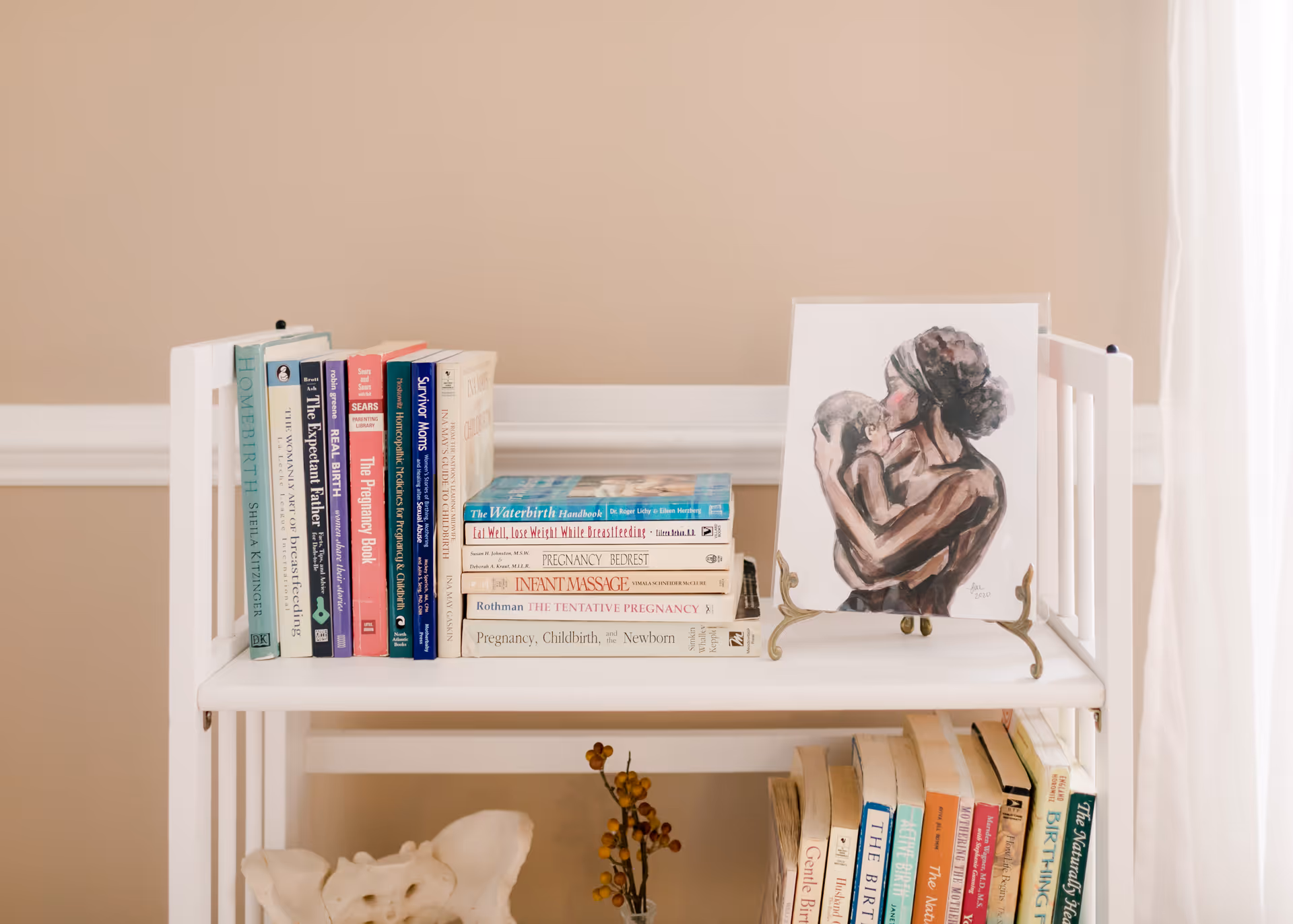 White bookshelf with stacked books and a framed mother-child drawing on a beige wall.